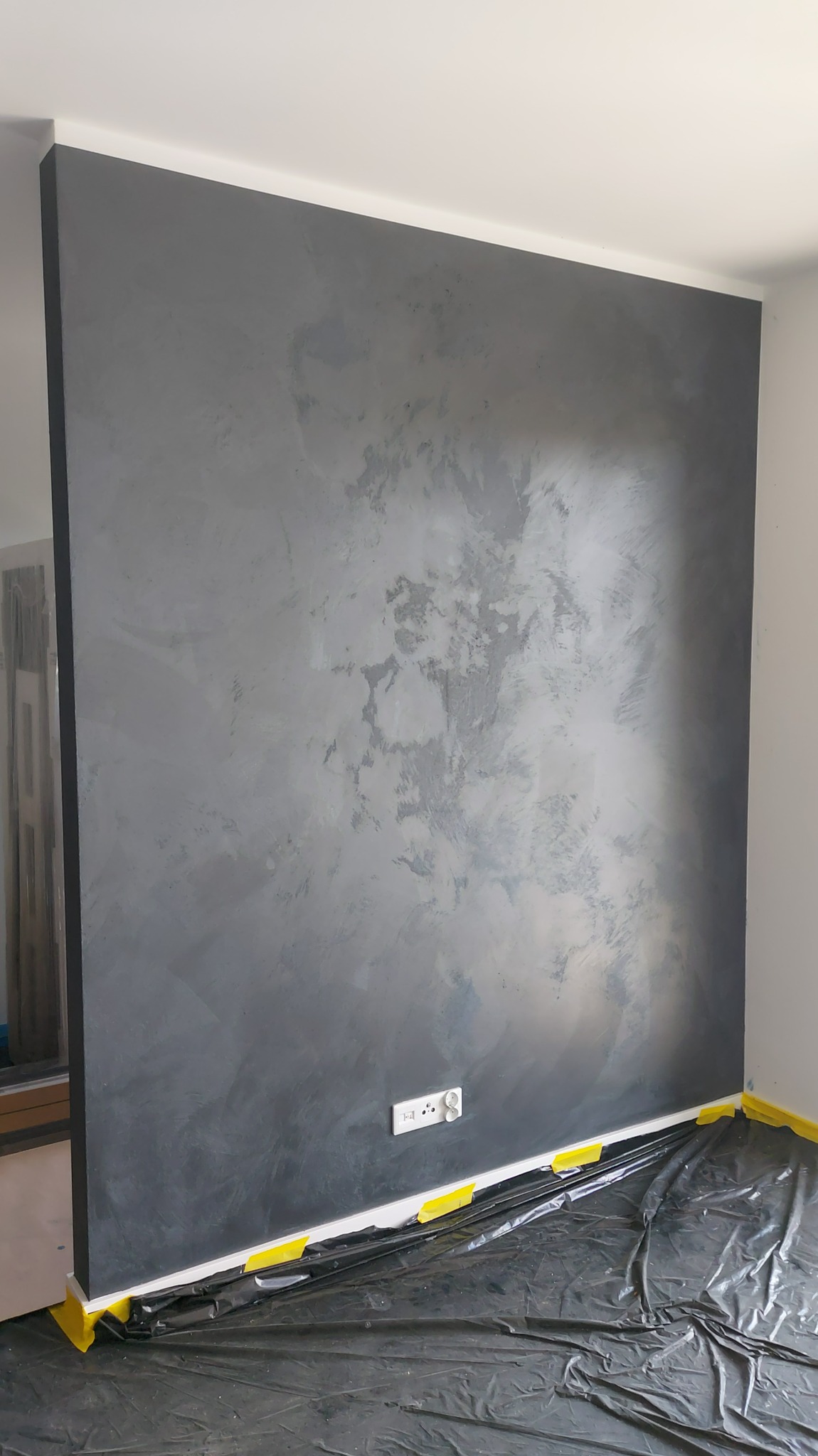 Ściana z efektem dekoracyjnym w odcieniach szarości, zabezpieczona folią malarską i taśmą. Widoczne gniazdko elektryczne. Charakterystyczna faktura ściany.