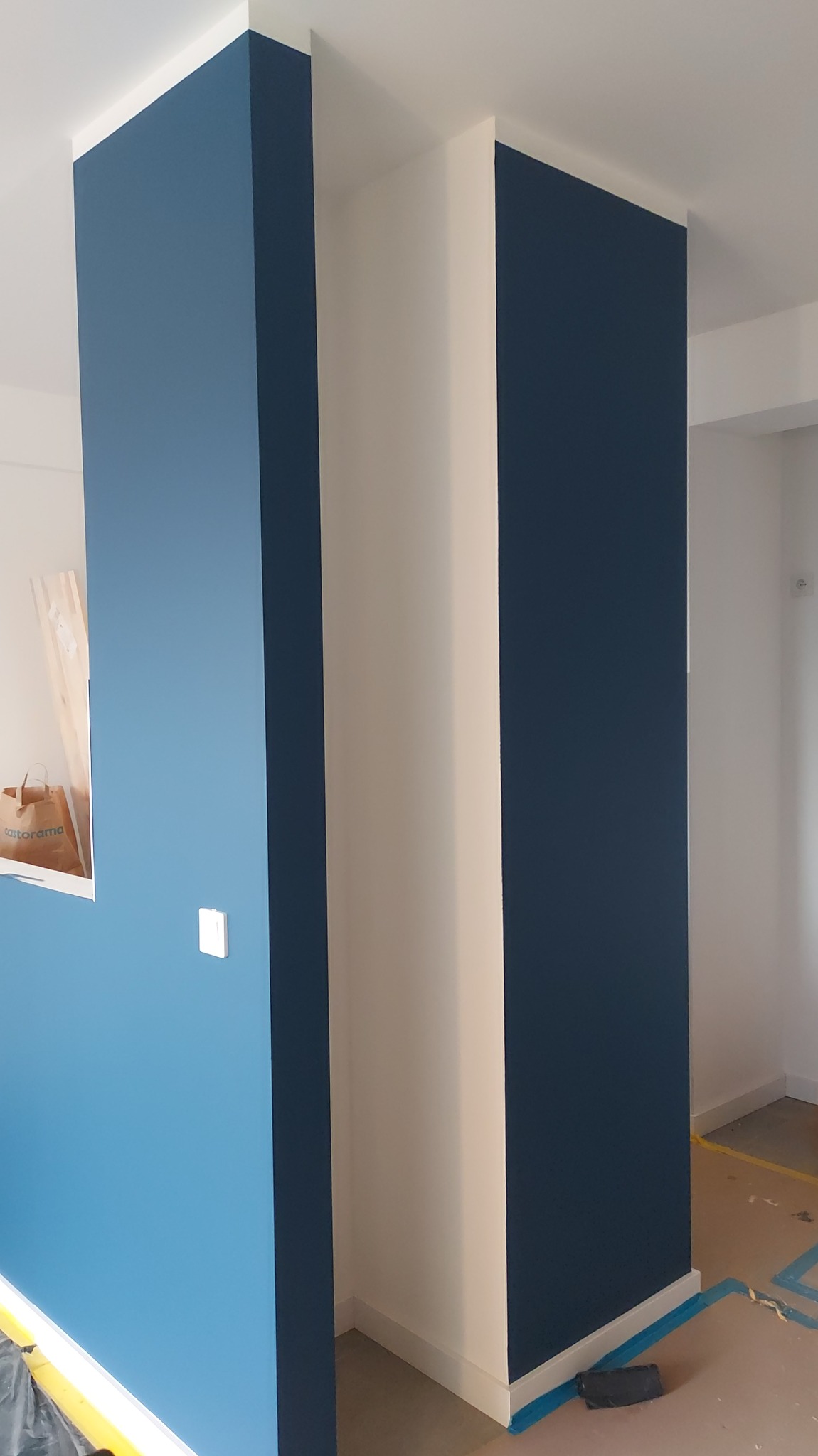 Świeżo pomalowane ściany w odcieniach błękitu i granatu, z białymi elementami wykończeniowymi. Widoczny fragment podłogi z zabezpieczeniem malarskim.