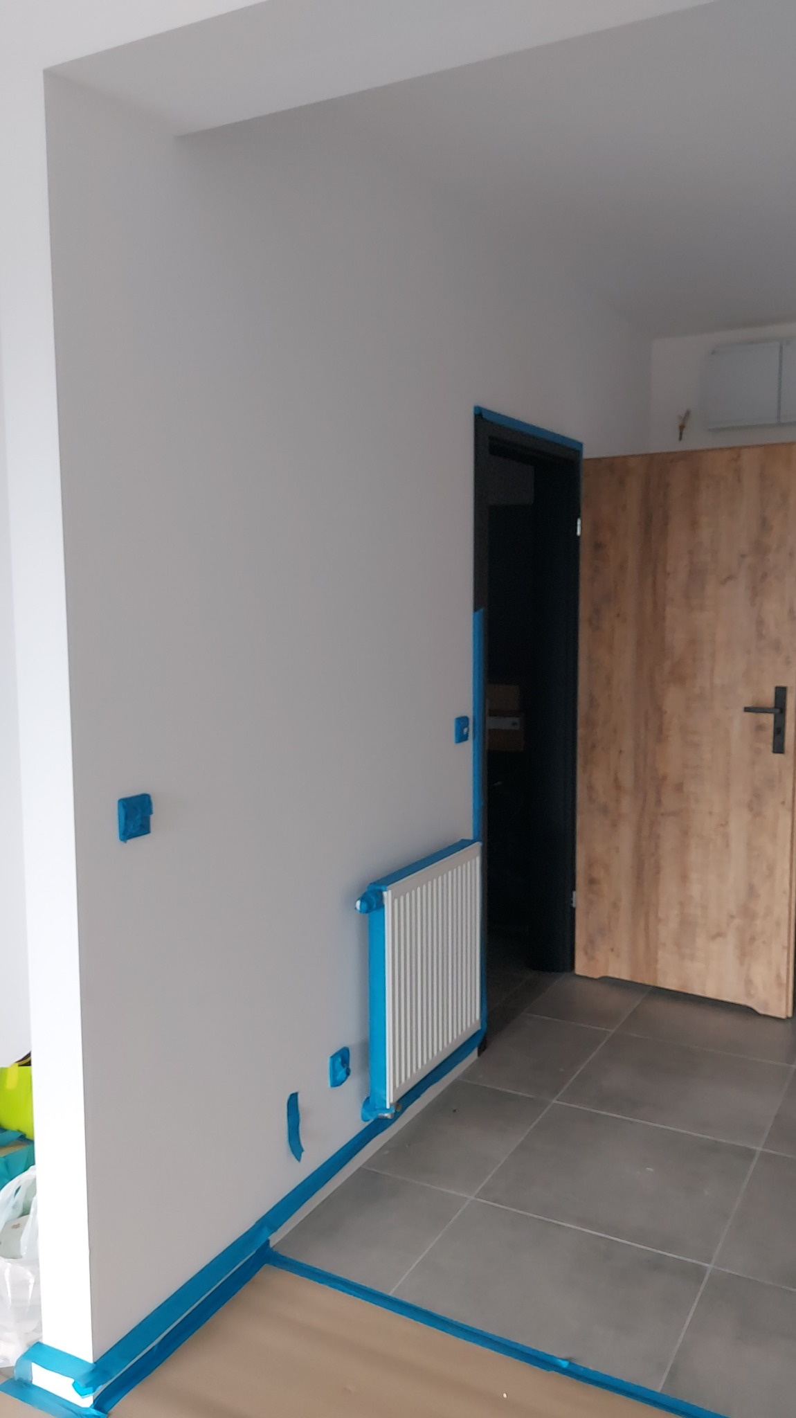 Przygotowane do malowania wnętrze z zabezpieczonymi taśmą grzejnikiem, listwami i gniazdkami. Ściany pomalowane na biało, widoczna drewniana stolarka drzwiowa.