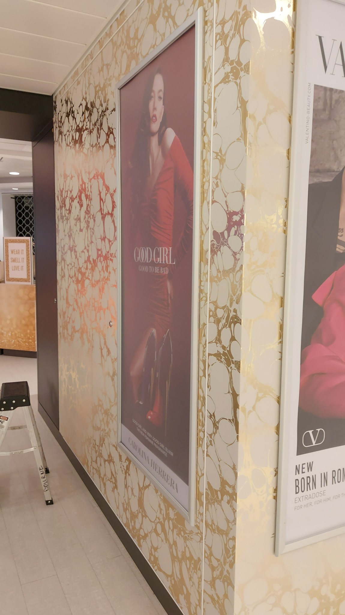 Ściana wykończona w marmurkowy wzór w odcieniach złota i bieli, z ramkami reklamowymi perfum, widoczne fragmenty drabiny malarskiej.