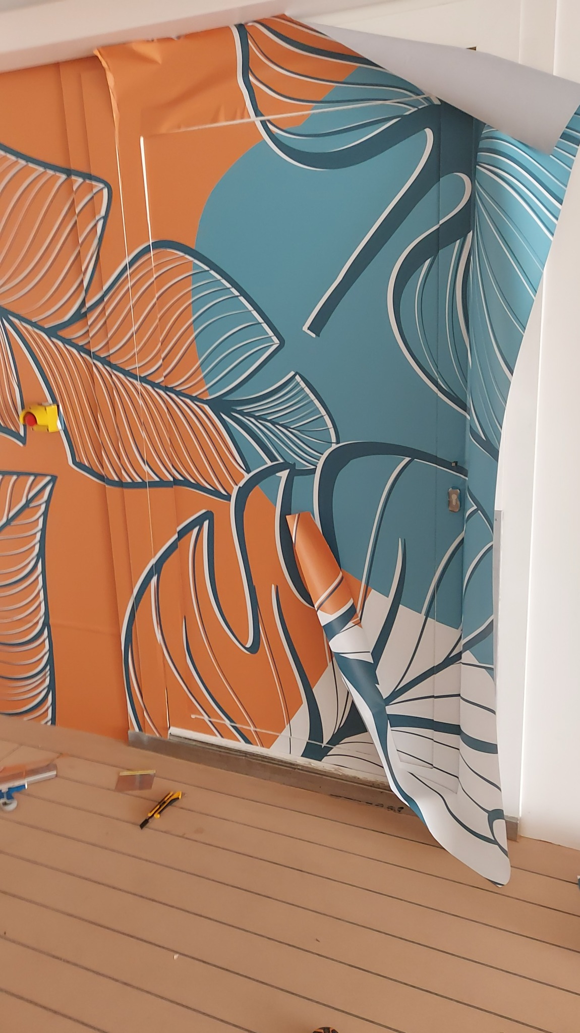 Naklejanie wzorzystej tapety na ścianę. Widoczne narzędzia i fragment odklejającej się tapety. Motyw roślinny, kolorystyka pomarańczowo-turkusowa.
