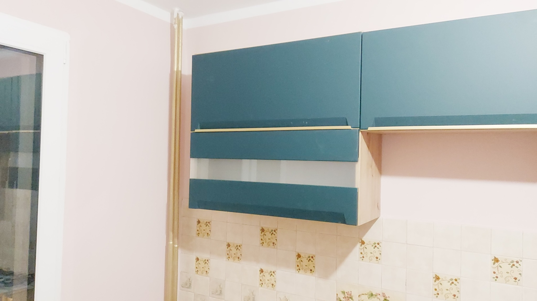 Turkusowe szafki wiszące z mlecznym szkłem w kuchni z kwiatowym wzorem na kafelkach. Widoczny fragment okna i pionowa listwa dekoracyjna.