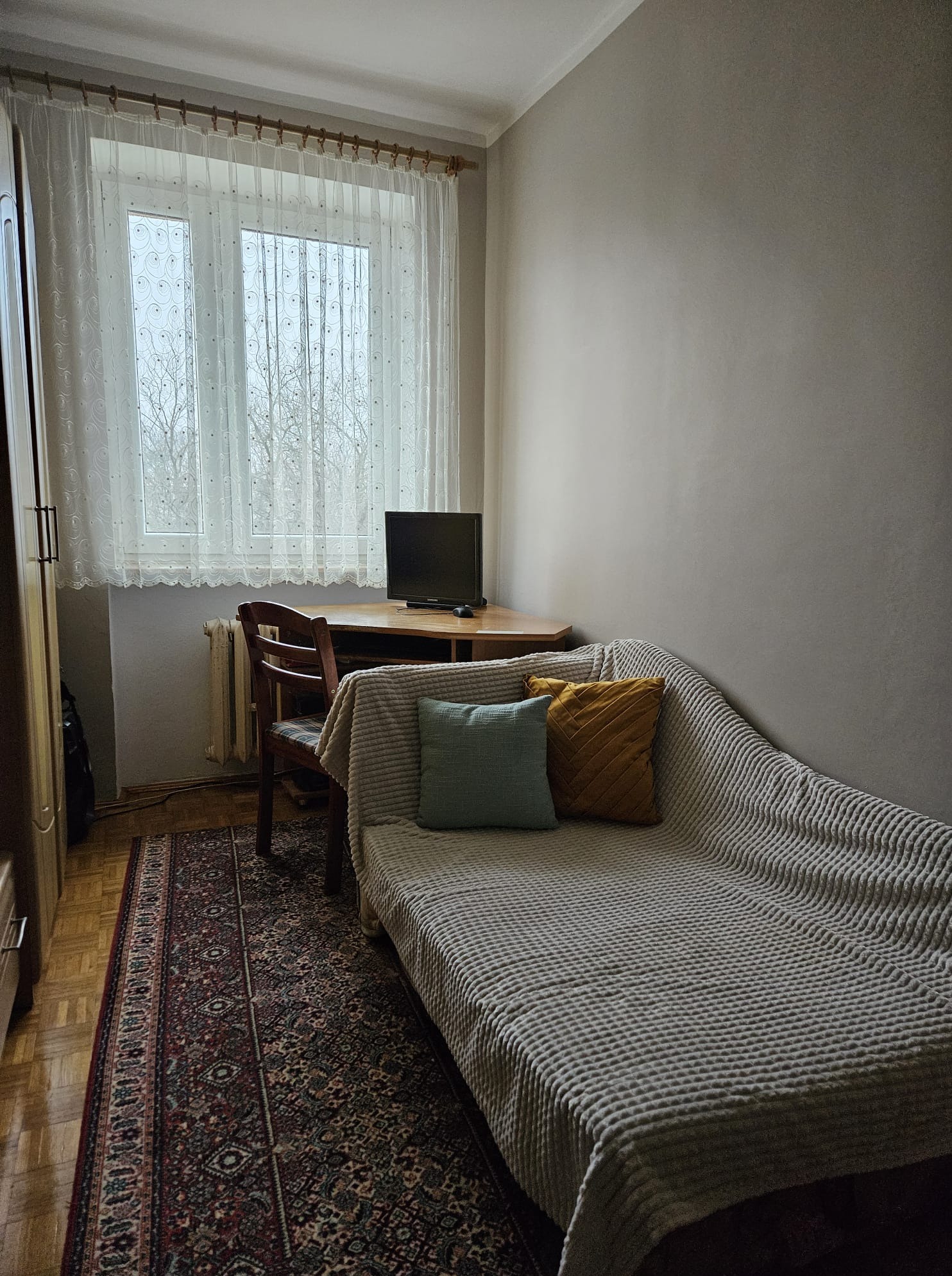 Jasne wnętrze pokoju z beżowymi ścianami, drewnianym biurkiem i tapczanem z dekoracyjnymi poduszkami. Widok na okno z firanką i dywan w orientalne wzory.