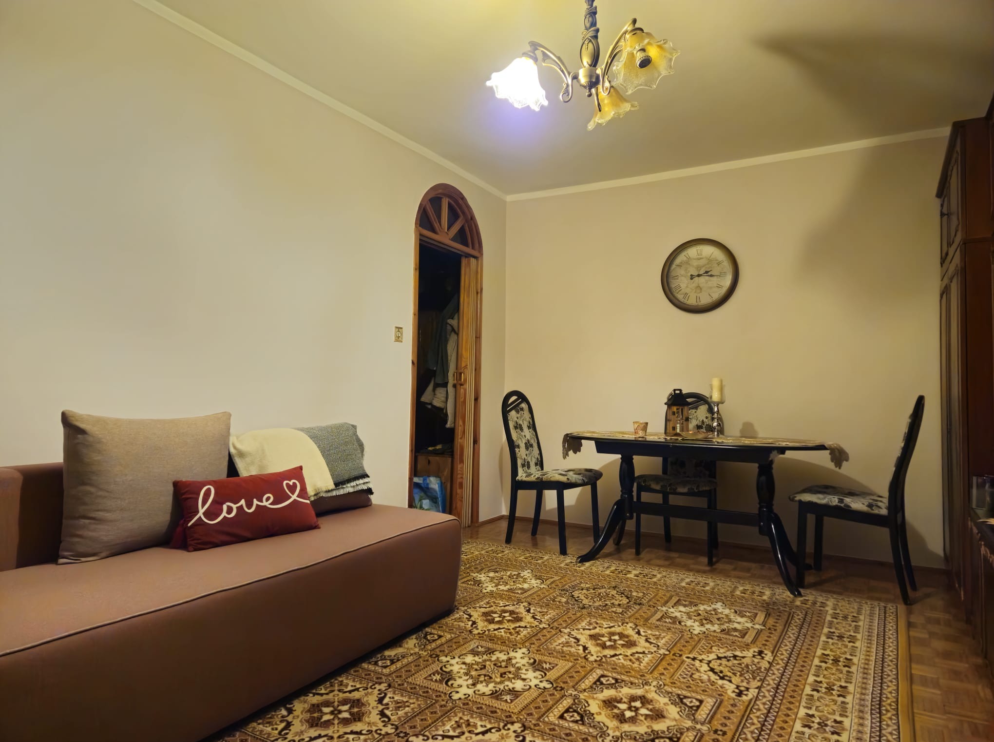 Przytulny salon z brązową sofą, wzorzystym dywanem i drewnianym stołem z krzesłami. Ciepłe oświetlenie i dekoracje tworzą domową atmosferę. Widoczny zegar na ścianie.