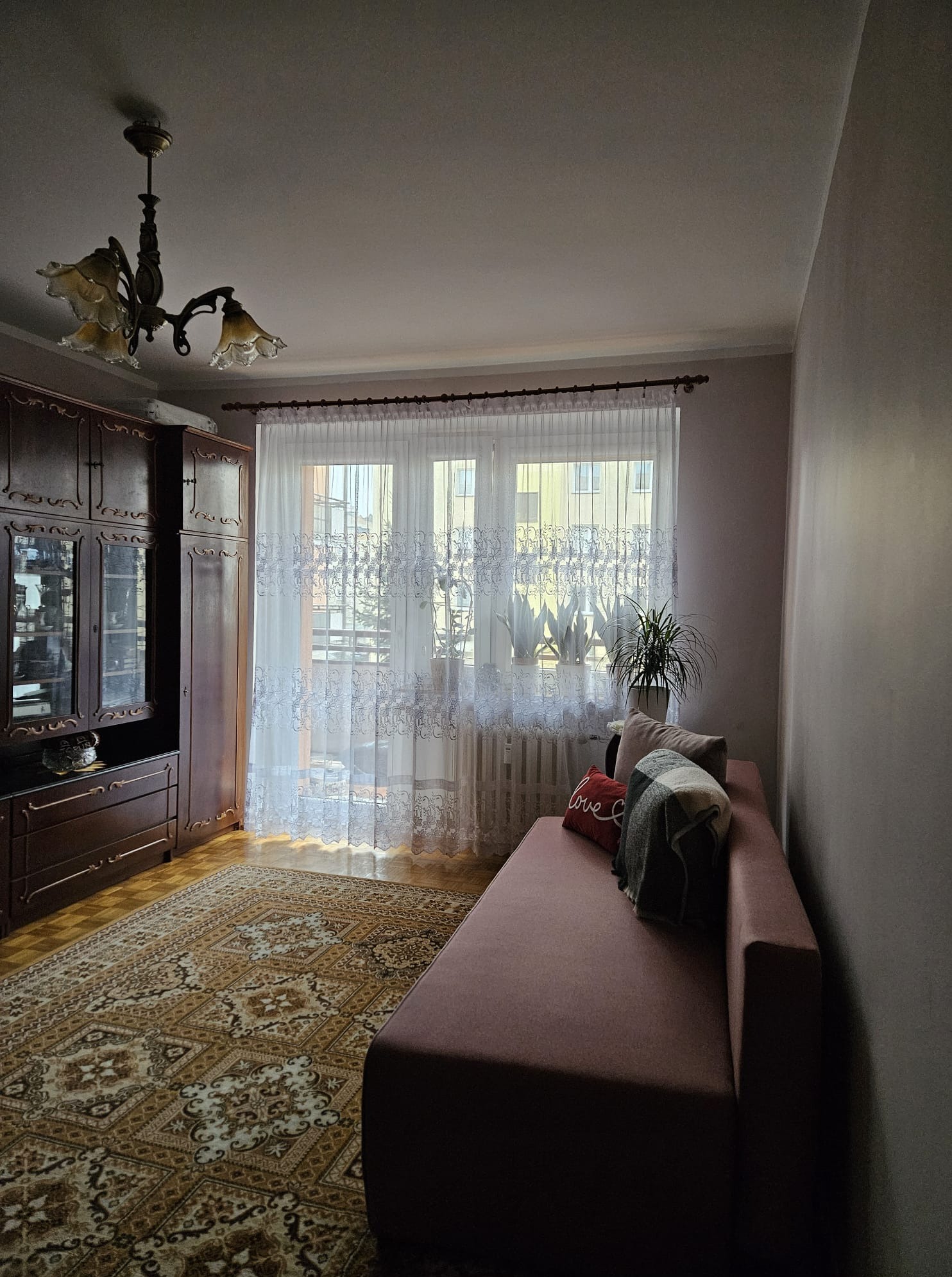 Pokój dzienny z brązowymi meblami, wzorzystym dywanem i różową sofą. Okno z firankami w tle. Widok na wnętrze po wykończeniu.