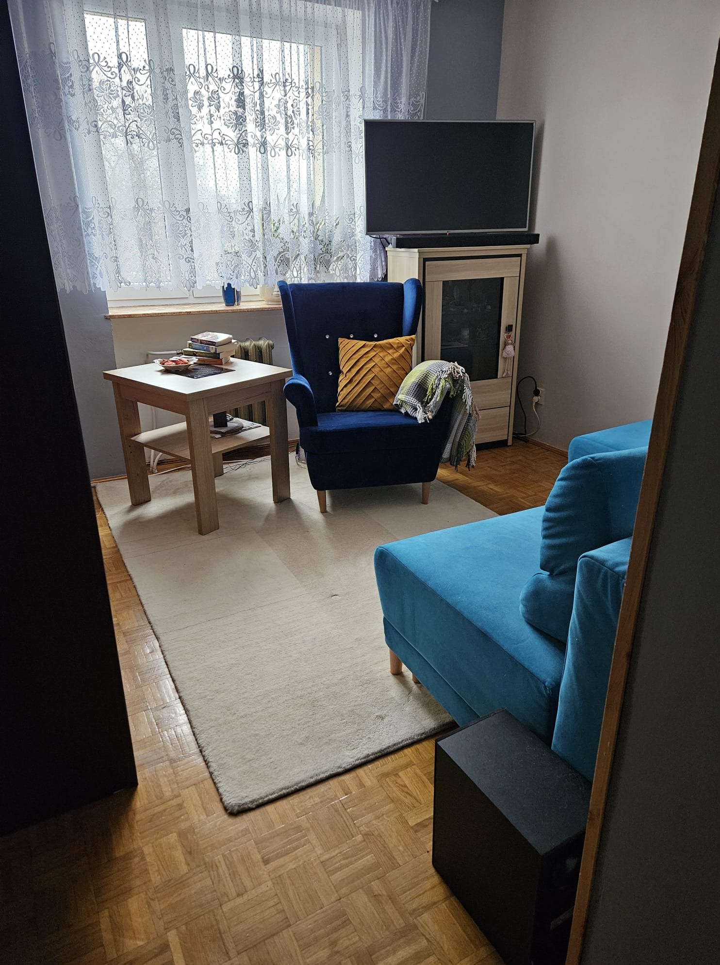 Aranżacja pokoju dziennego: turkusowa sofa, granatowy fotel z żółtą poduszką, drewniany stolik i dywan na parkiecie. Nowoczesne wnętrze po odświeżeniu.