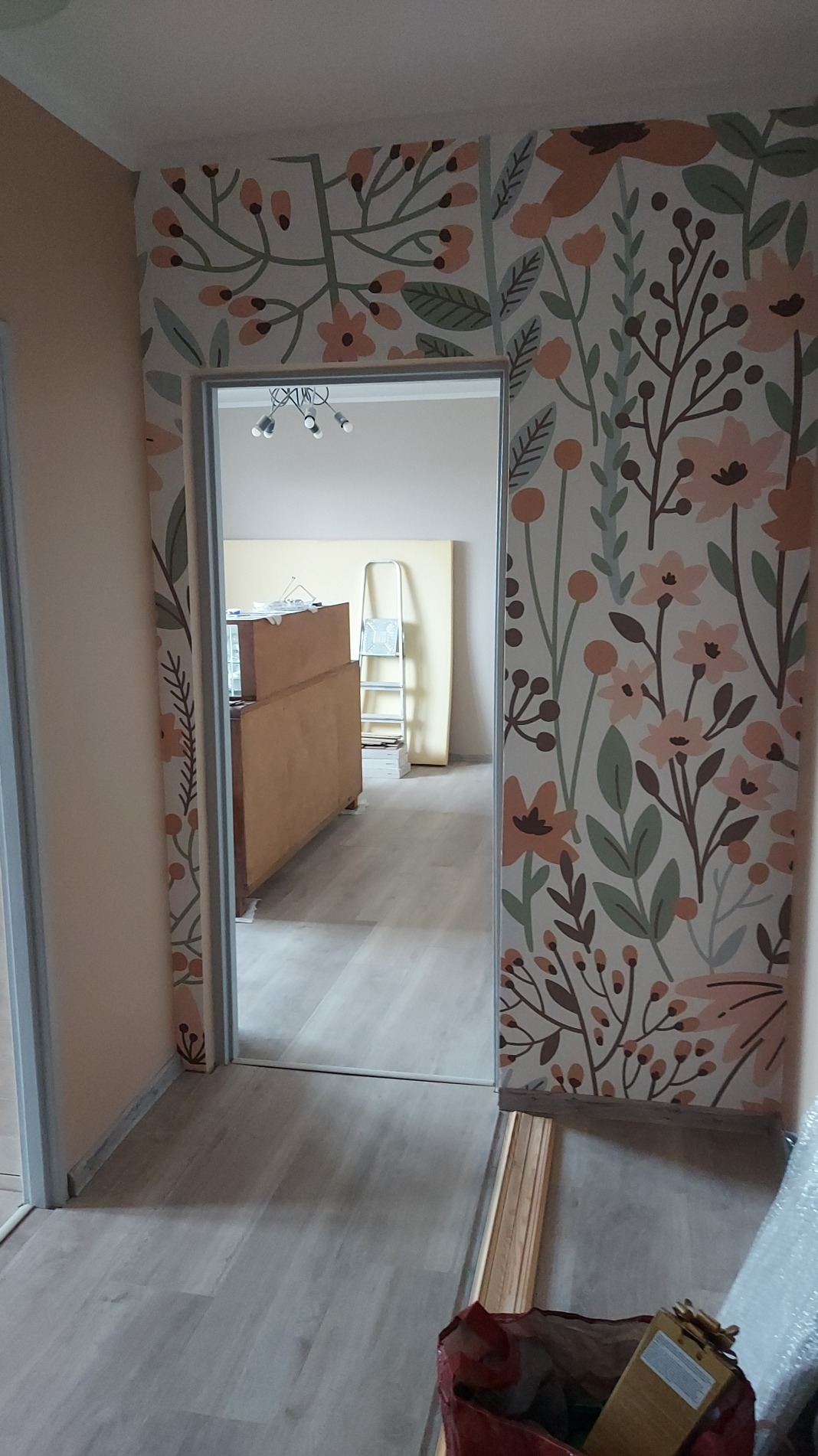 Korytarz po remoncie: kwiatowy wzór tapety na ścianach, jasne panele podłogowe, widok na sąsiednie pomieszczenie z meblami i drabiną.