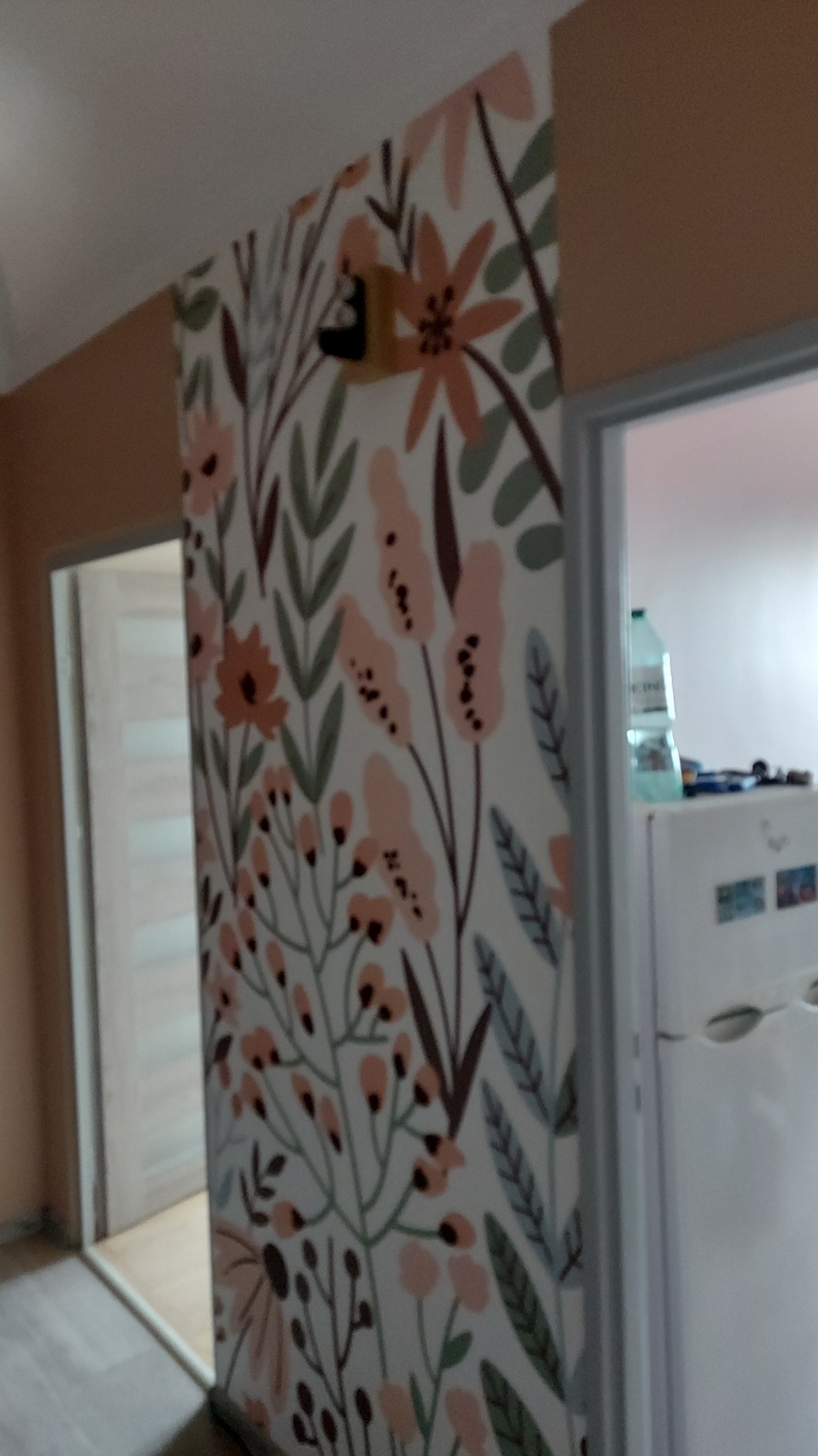 Ściana z dekoracyjną tapetą w kwiaty w mieszkaniu, widoczna z perspektywy narożnika pokoju, z fragmentem drzwi i sprzętu AGD w tle.