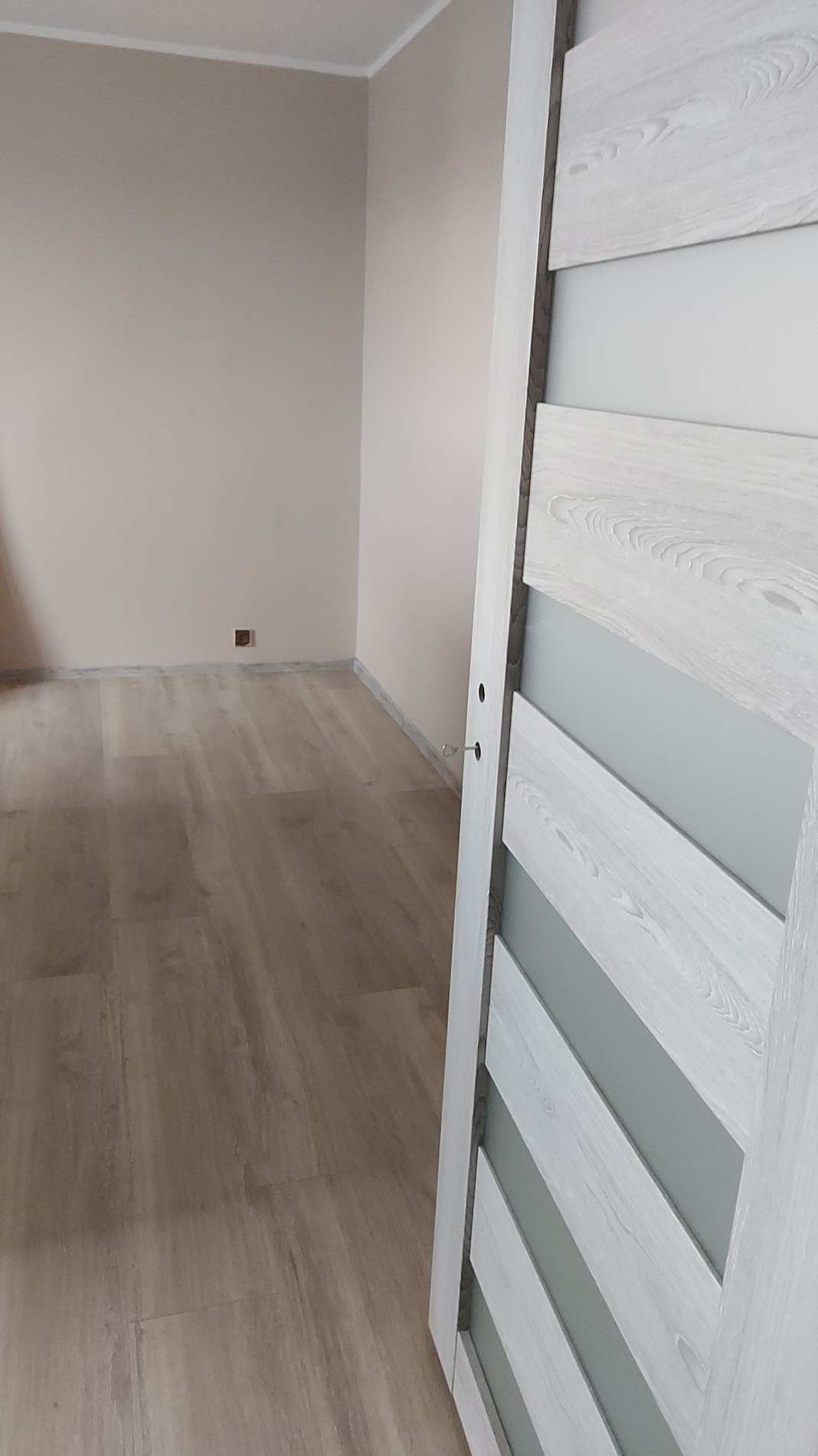 Jasne panele podłogowe i beżowe ściany w pokoju po remoncie. W tle widoczny fragment białych drzwi z przeszkleniami. Minimalistyczne wykończenie wnętrza.