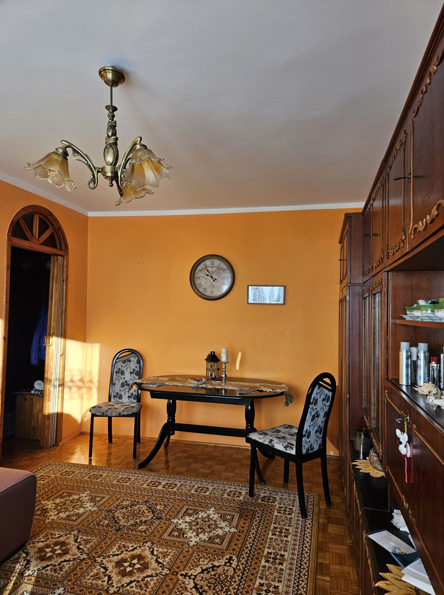 Pokój z pomarańczowymi ścianami, drewnianą podłogą i dywanem w orientalnym stylu. Widoczne meble: stół, krzesła, kredens. Na ścianie zegar i obrazek.