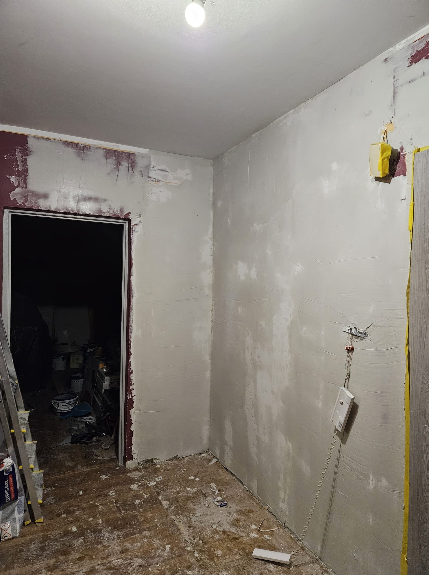 Pokój w trakcie gładziowania ścian, widoczne ślady po wcześniejszym malowaniu, zabezpieczone elementy, drabina i narzędzia na podłodze. Wnętrze w stanie surowym.