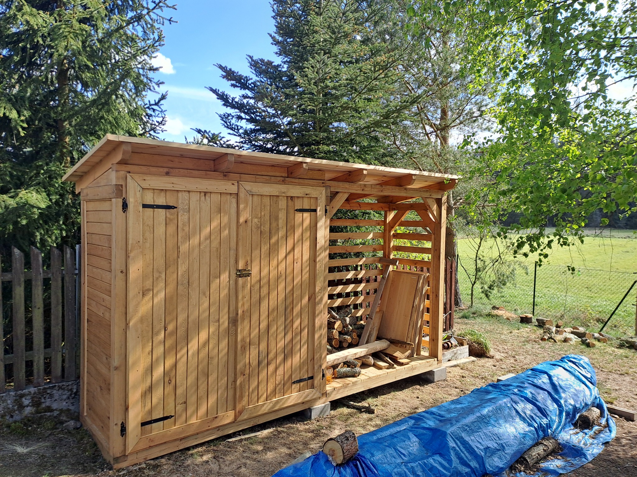 Drewniana wiata na drewno z zamkniętą częścią i otwartą kratownicą, na tle zieleni i błękitnego nieba, przykryta plandeką.