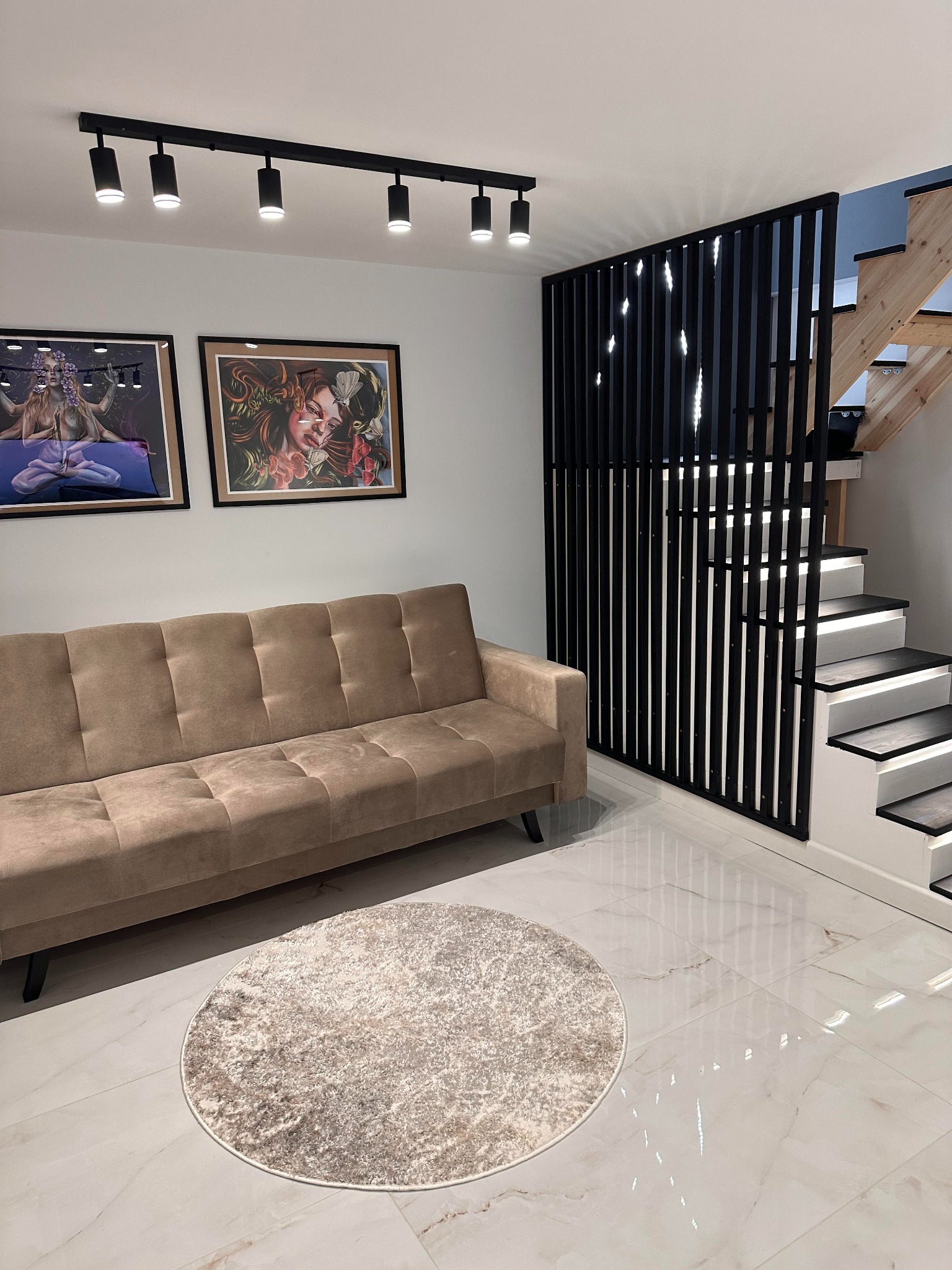 Nowoczesne wnętrze: beżowa sofa, okrągły dywan, białe płytki, czarna ażurowa ścianka działowa i oświetlone schody. Na ścianie wiszą dwa obrazy.