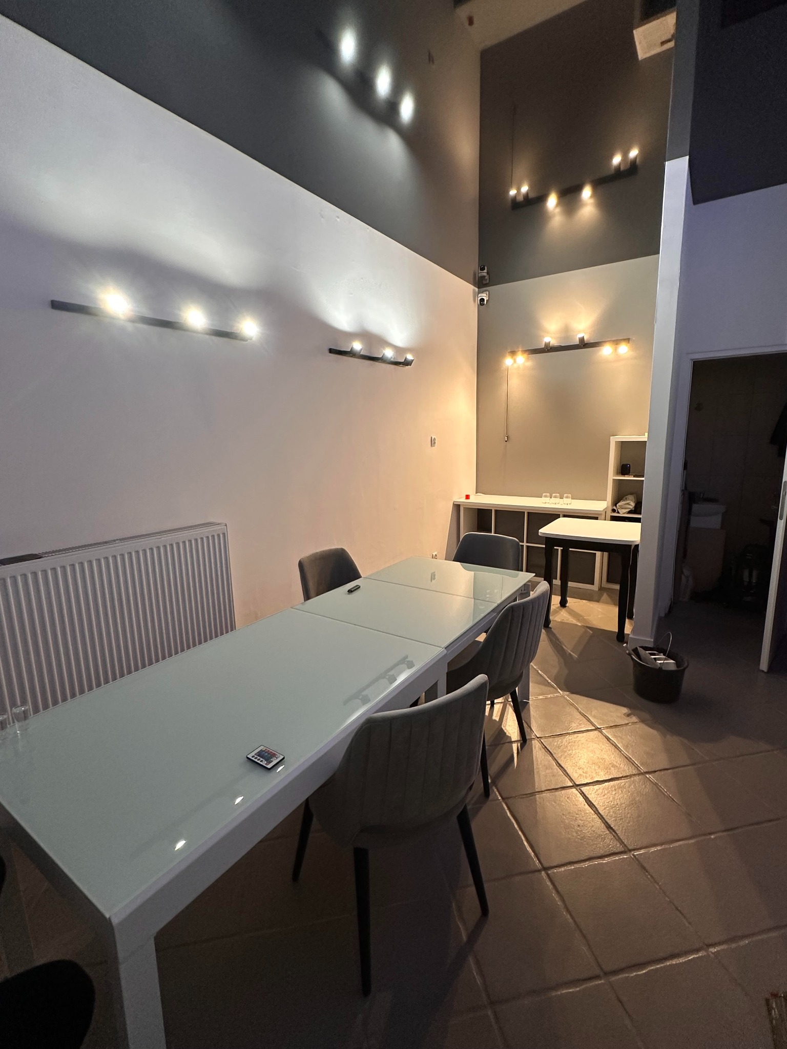 Jasne biuro po odświeżeniu: białe ściany z rzędami lamp, szary sufit, szklany stół z krzesłami. Widoczny grzejnik i fragment podłogi z płytek. Nowoczesny design.