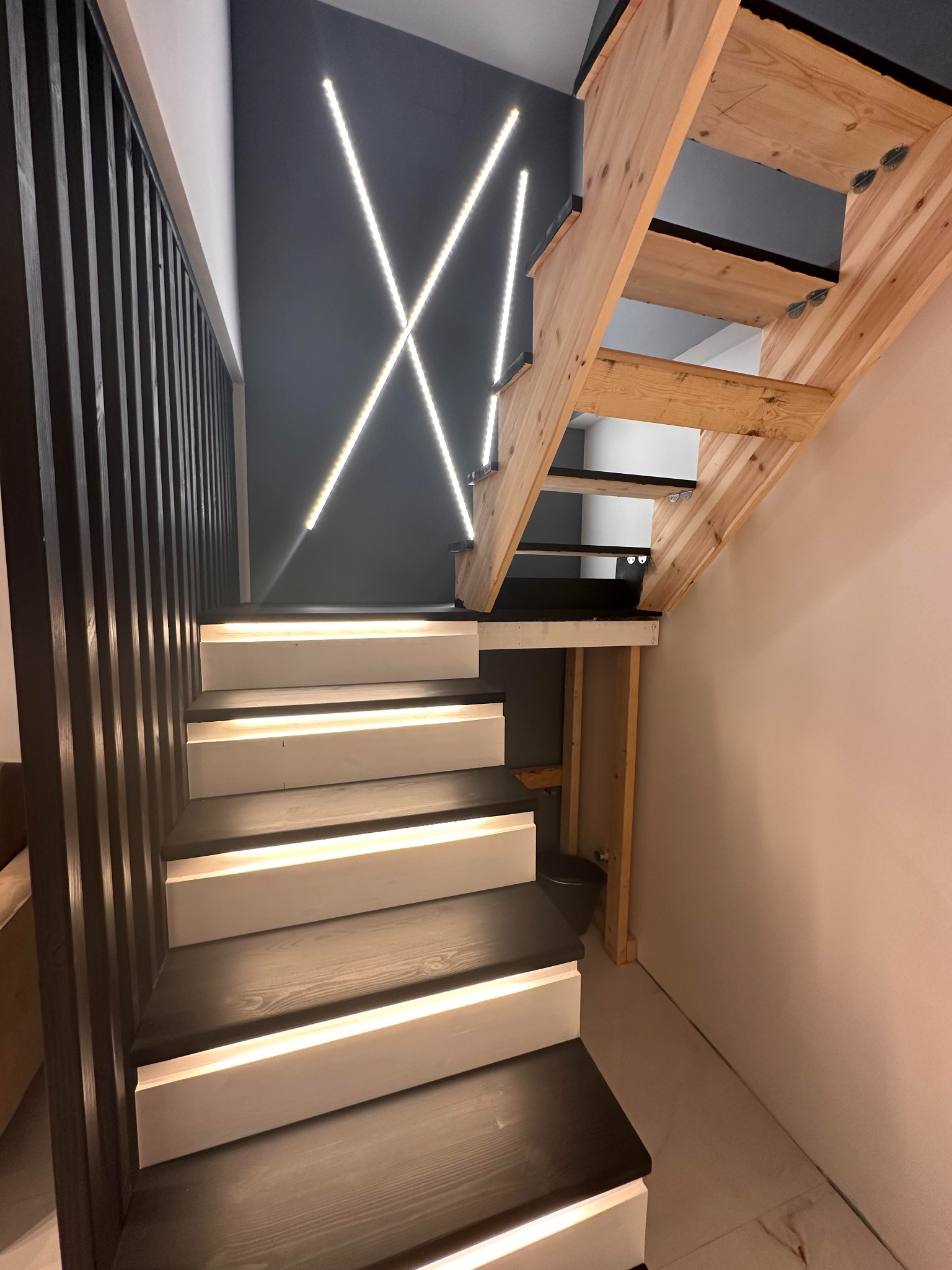 Nowoczesne schody z podświetleniem LED na ścianie, czarne stopnie i białe podstopnice, drewniane barierki, surowa konstrukcja schodów na piętro.