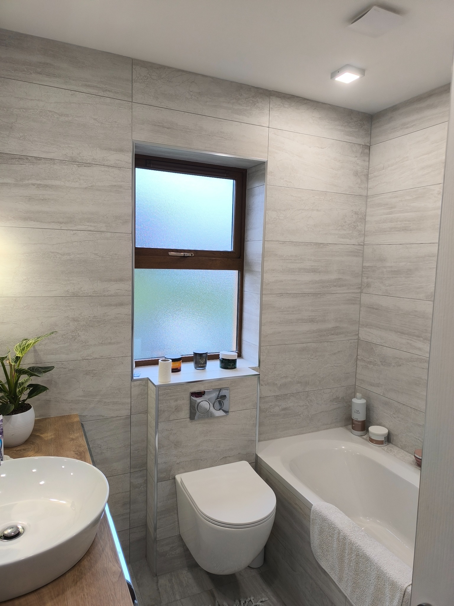 Jasna łazienka z wanną, umywalką i WC w zabudowie. Ściany wyłożone płytkami imitującymi drewno. Okno z brązową ramą. Minimalistyczny styl.