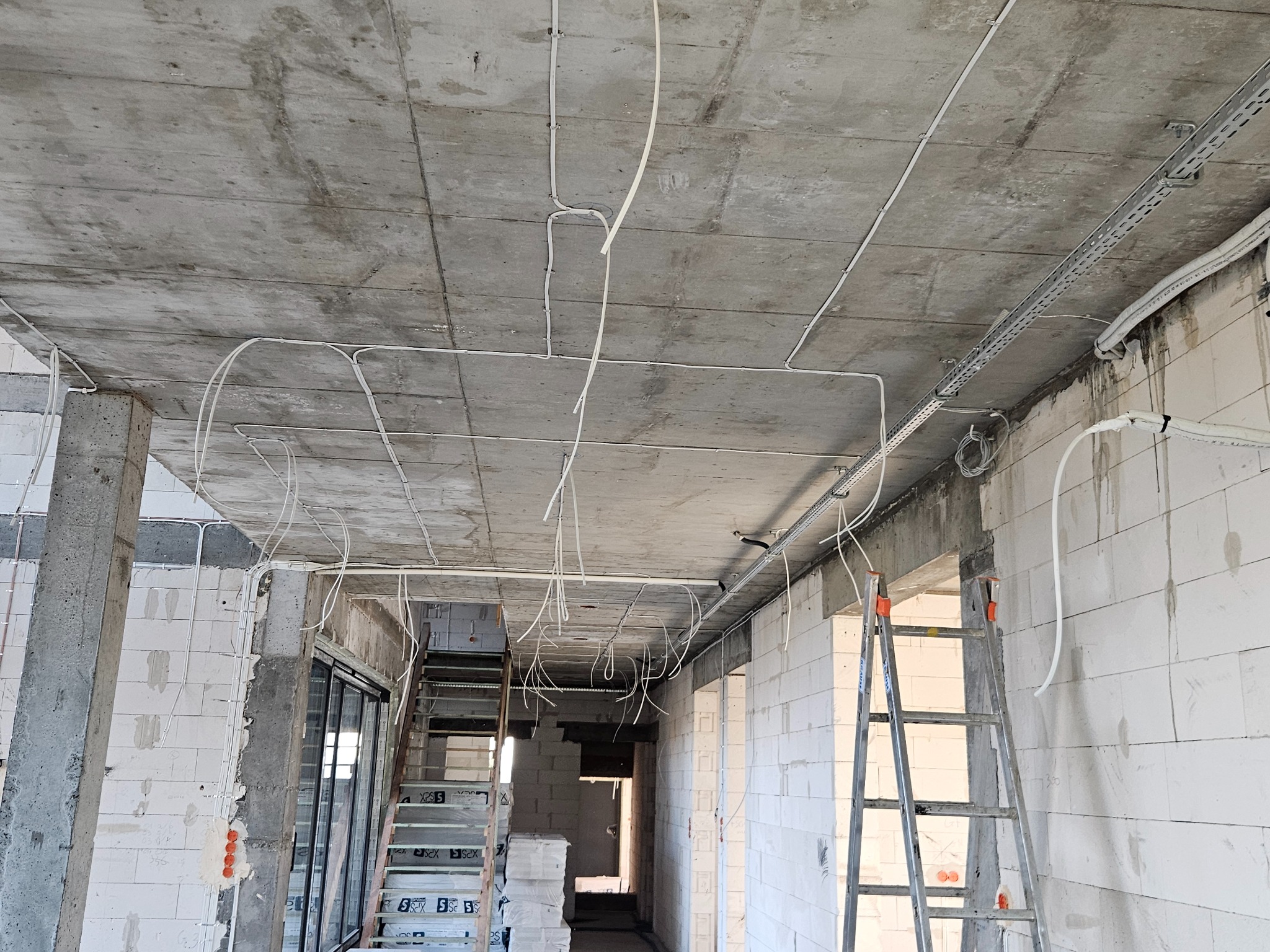 Instalacja elektryczna w surowym budynku: kable na suficie, drabina, ściany z betonu komórkowego. Widoczne przewody w korytach kablowych i luźno zwisające.