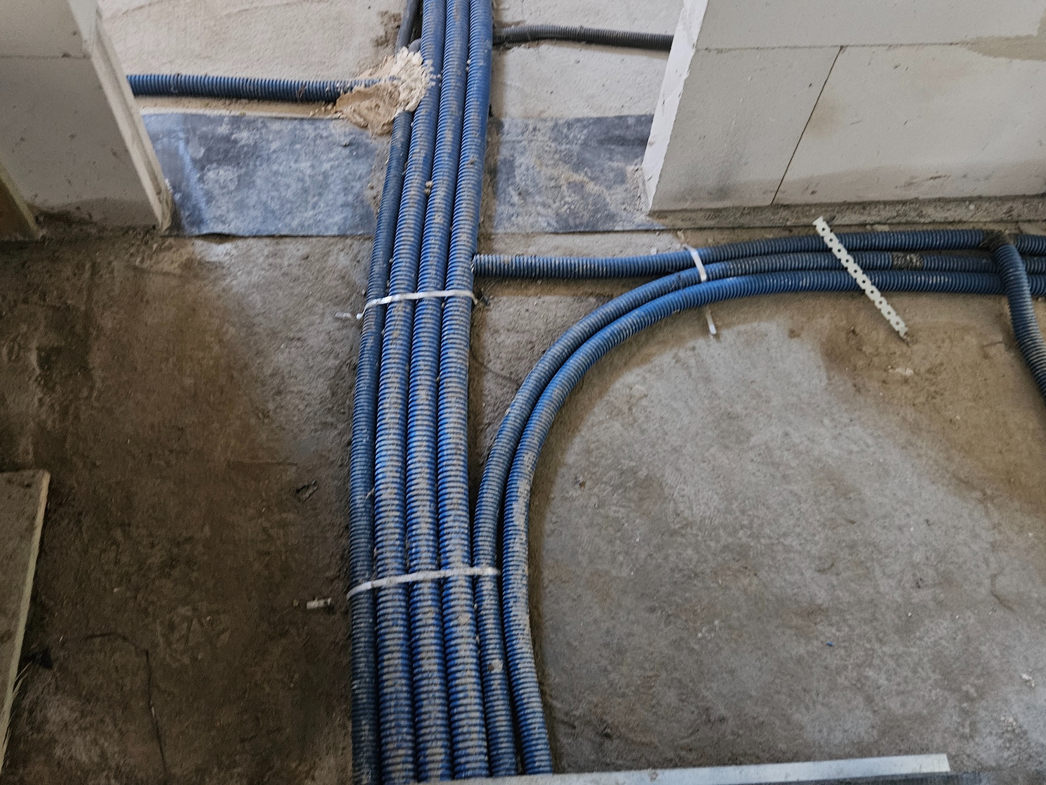Niebieskie rury karbowane, częściowo przymocowane, ułożone na surowym podłożu w trakcie prac instalacyjnych wewnątrz budynku. Widoczne elementy konstrukcyjne.