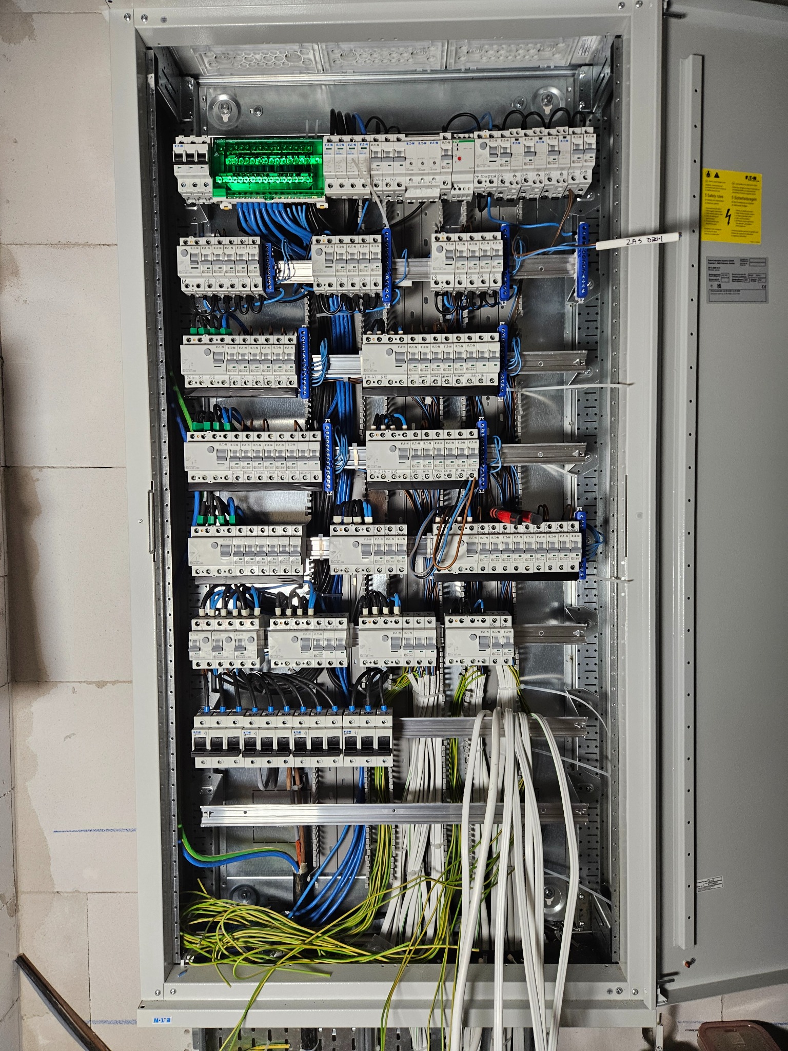 Otwarta szafa elektryczna z licznymi zabezpieczeniami i przewodami w kolorach: niebieskim, zielono-żółtym i białym, zamontowana na ścianie z bloczków.