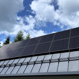 MTech Instalacje S.C. - Czarny dach z panelami fotowoltaicznymi w słoneczny dzień. Widoczne mocowania paneli. W tle drzewa i błękitne niebo z chmurami.