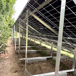 MTech Instalacje S.C. - Widok od spodu na panele fotowoltaiczne zamontowane na metalowej konstrukcji, z zielenią w tle. Perspektywa z dołu podkreśla regularność i skalę instalacji.