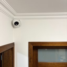 MTech Instalacje S.C. - Biała kamera kopułkowa zamontowana w rogu pokoju z dekoracyjną listwą sufitową i fragmentami drewnianych futryn drzwiowych.