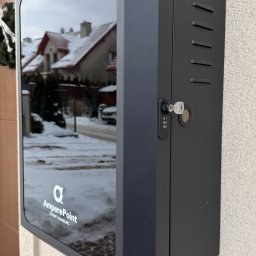MTech Instalacje S.C. - Zamontowana stacja ładowania pojazdów elektrycznych AmperePoint na ścianie budynku w Brzesku. Widoczny zamek i szyba z odbiciem otoczenia.