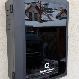 MTech Instalacje S.C. - Szara stacja ładowania samochodów elektrycznych AmperPoint zamontowana na elewacji budynku. W odbiciu widoczny dom z panelami słonecznymi i zaparkowany samochód.