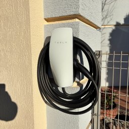 MTech Instalacje S.C. - Biała ładowarka Tesla na szarej ścianie budynku, z czarnym kablem zwiniętym obok. Widoczne fragmenty elewacji w odcieniach beżu i szarości oraz metalowe ogrodzenie.