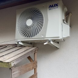 MTech Instalacje S.C. - Biała jednostka zewnętrzna klimatyzacji AUX DC Inverter zamontowana na ścianie budynku, widoczna z boku, z drewnianą konstrukcją i stosem drewna w tle.