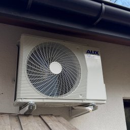 MTech Instalacje S.C. - Zamontowana jednostka zewnętrzna klimatyzacji AUX DC Inverter na elewacji budynku, widoczna z dołu, z fragmentem drewnianej konstrukcji w kadrze.