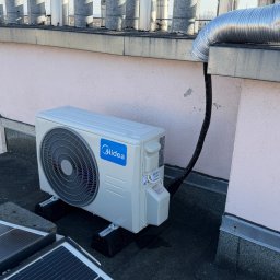 MTech Instalacje S.C. - Klimatyzator Midea na dachu obok paneli słonecznych i kominów wentylacyjnych. Widoczny przewód odprowadzający. Jasne niebo w tle. Instalacja na dachu budynku.