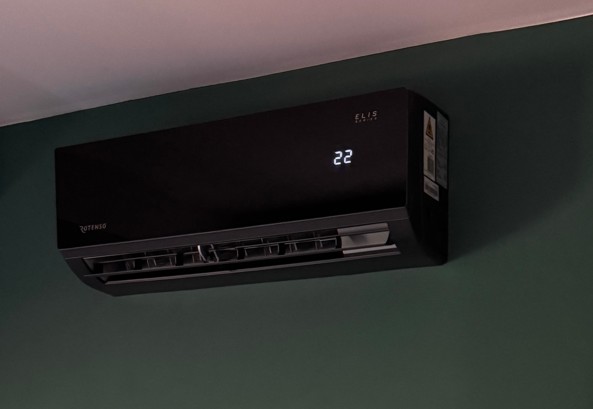Czarny klimatyzator Rotenso Elis Series, zamontowany na zielonej ścianie, wyświetlający temperaturę 22 stopnie. Widoczne logo i ostrzeżenia na boku urządzenia.