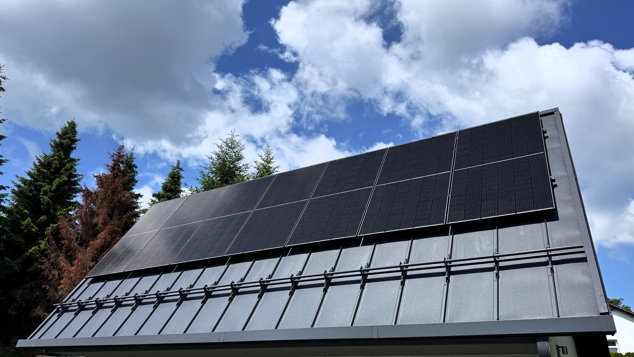 Czarny dach z panelami fotowoltaicznymi w słoneczny dzień. Widoczne mocowania paneli. W tle drzewa i błękitne niebo z chmurami.