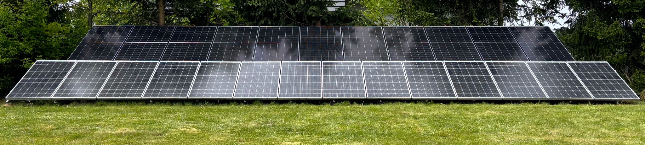 Dwa rzędy paneli fotowoltaicznych na trawniku, z drzewami w tle. Panele ułożone poziomo, tworząc długą instalację. Widoczne mocowania paneli.