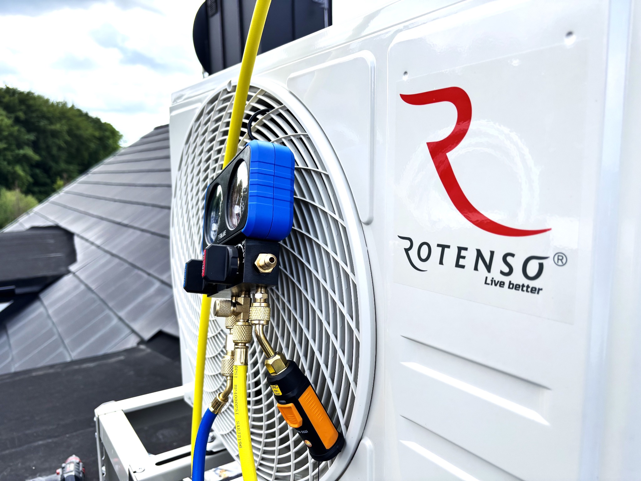 Biała jednostka zewnętrzna klimatyzacji Rotenso na dachu, podłączona do niebiesko-żółtego zestawu manometrów serwisowych, z widocznym fragmentem dachu i zieleni w tle.