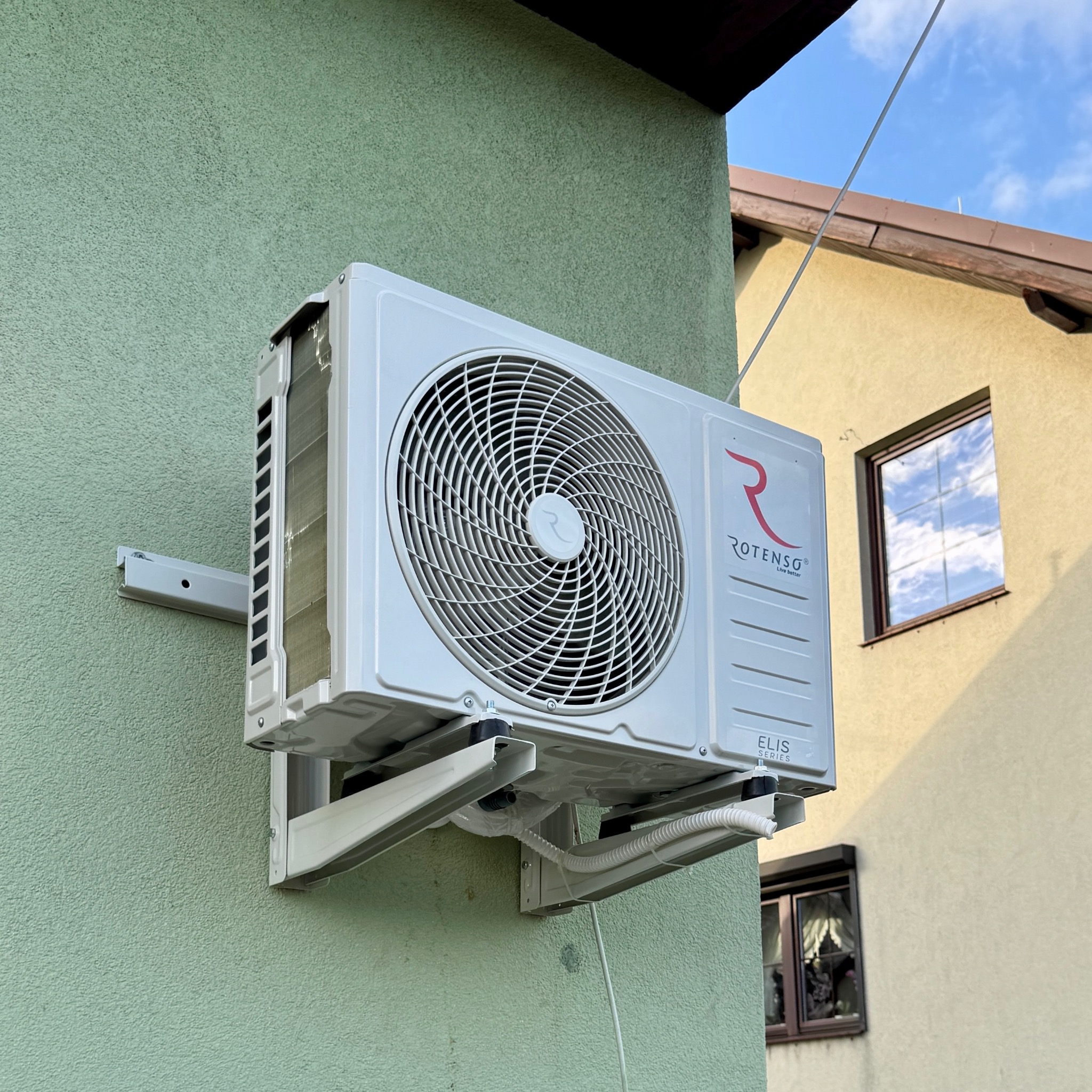 Biała jednostka zewnętrzna klimatyzacji Rotenso Elis Series zamontowana na zielonej elewacji budynku, widok z dołu, błękitne niebo w tle.