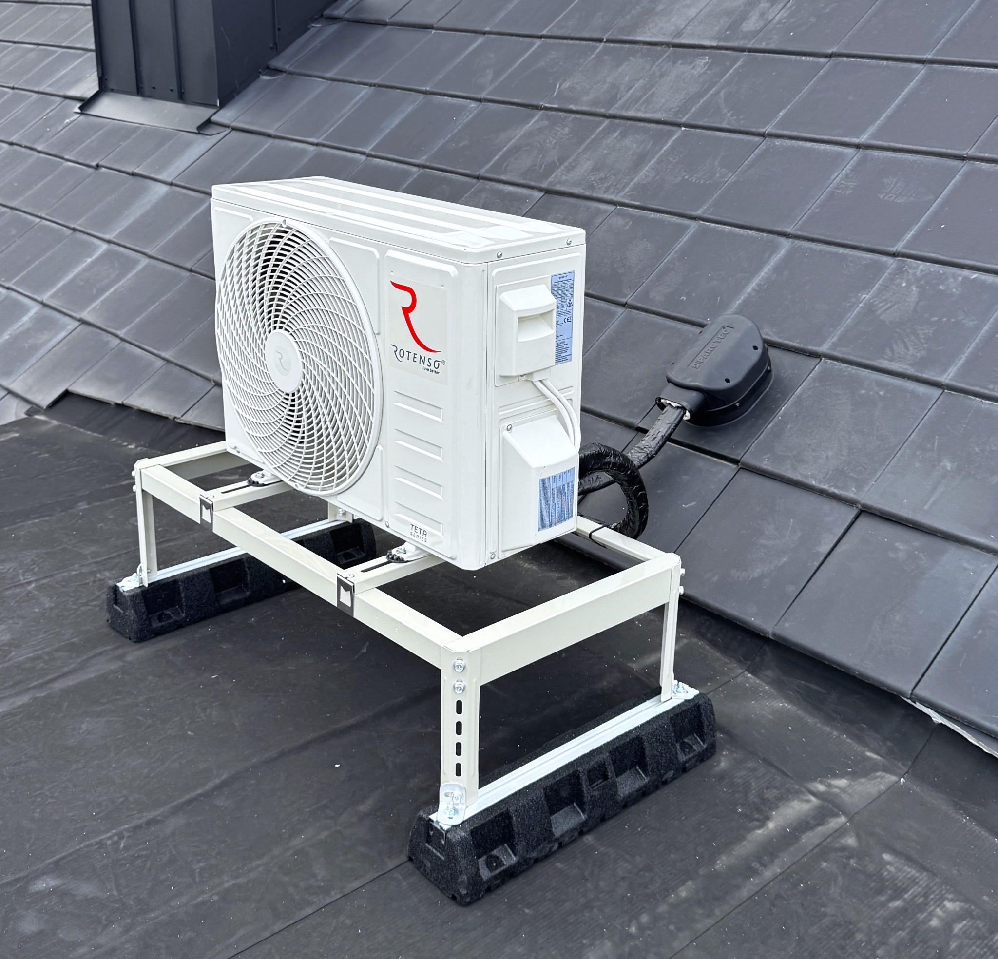 Biała jednostka zewnętrzna klimatyzacji Rotenso na dachu z szarej dachówki, zamontowana na metalowej konstrukcji z czarnymi podkładkami antywibracyjnymi.
