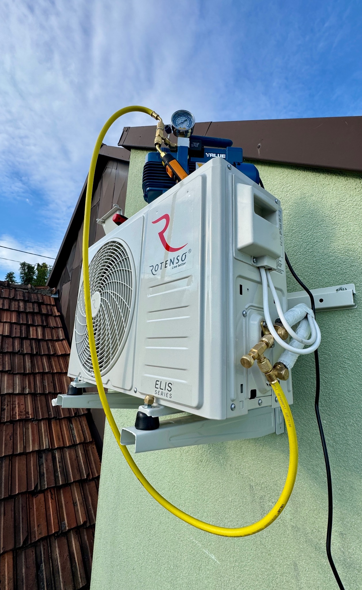 Biała jednostka klimatyzacji Rotenso Elis Series zamontowana na zielonej ścianie budynku, z podłączonymi przewodami i pompą próżniową. Widoczne dachówki i błękitne niebo.