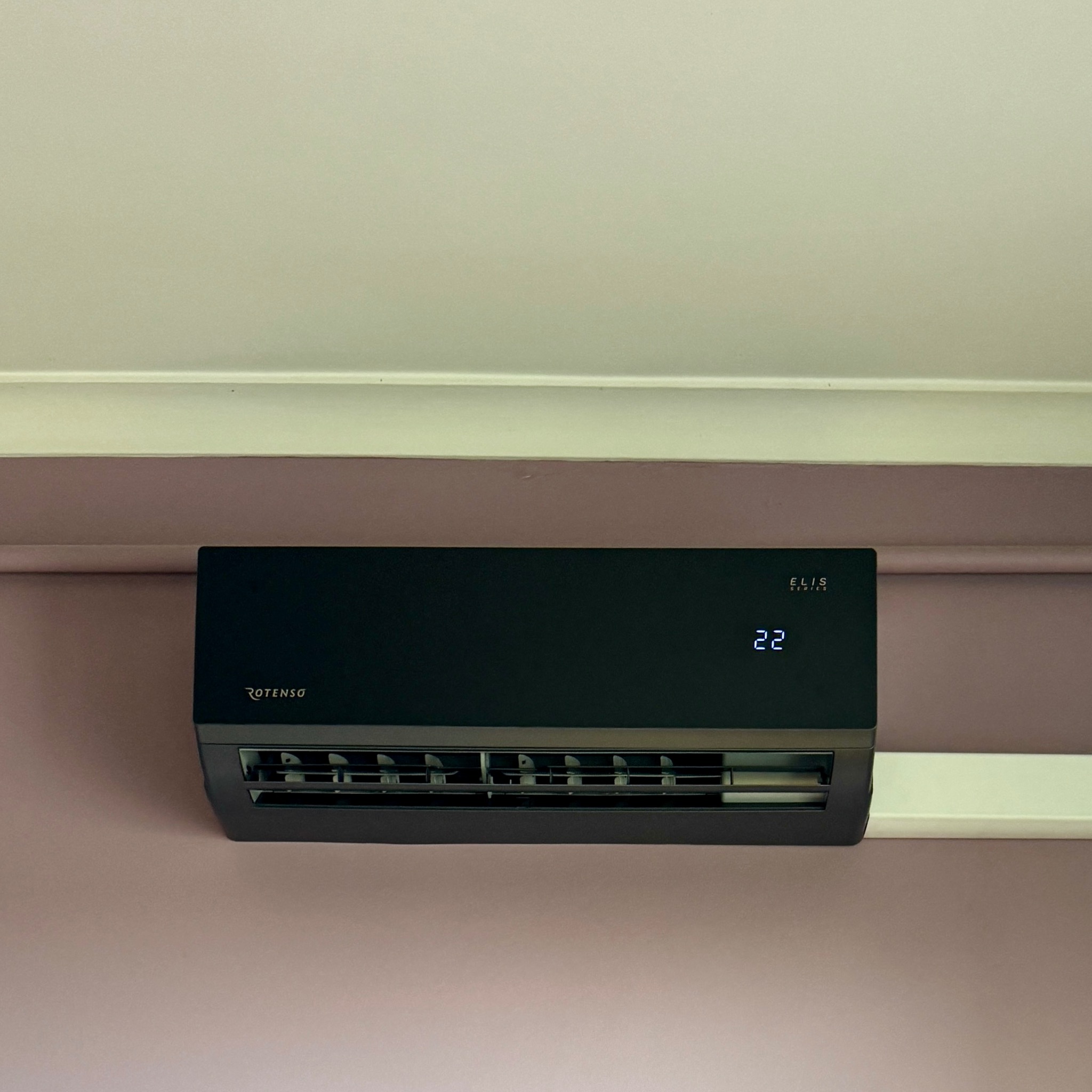 Czarny klimatyzator Rotenso Elis Series z wyświetlaczem temperatury 22, zamontowany na liliowej ścianie pod sufitem. Widoczne żaluzje wylotu powietrza.