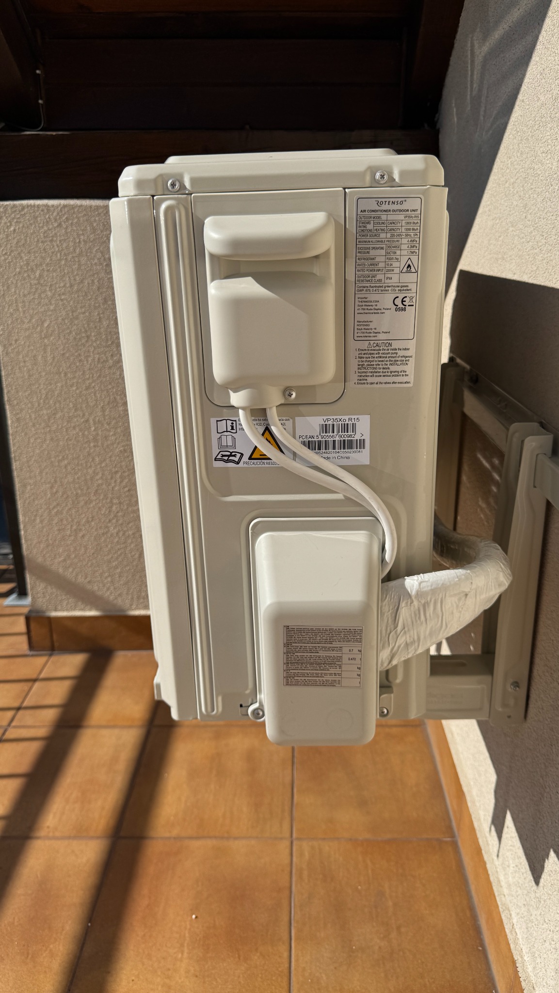 Zewnętrzna jednostka klimatyzacji Rotenso, zamontowana na ścianie budynku, widoczna z tyłu, z widocznymi etykietami informacyjnymi i przewodami.