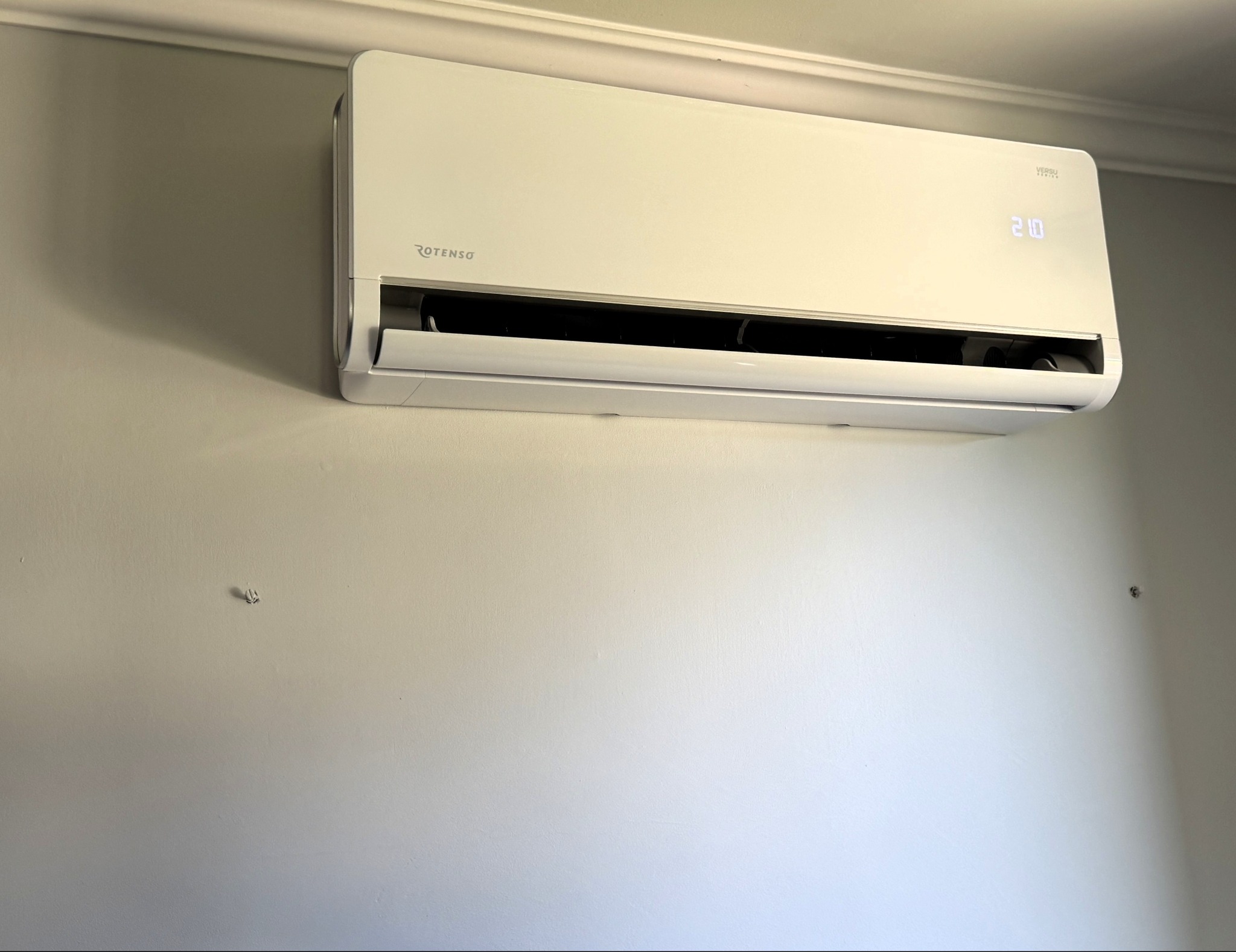 Biała klimatyzacja Rotenso Versu Series zamontowana na ścianie. Widoczny wyświetlacz z temperaturą 21 stopni. Minimalistyczny design urządzenia.