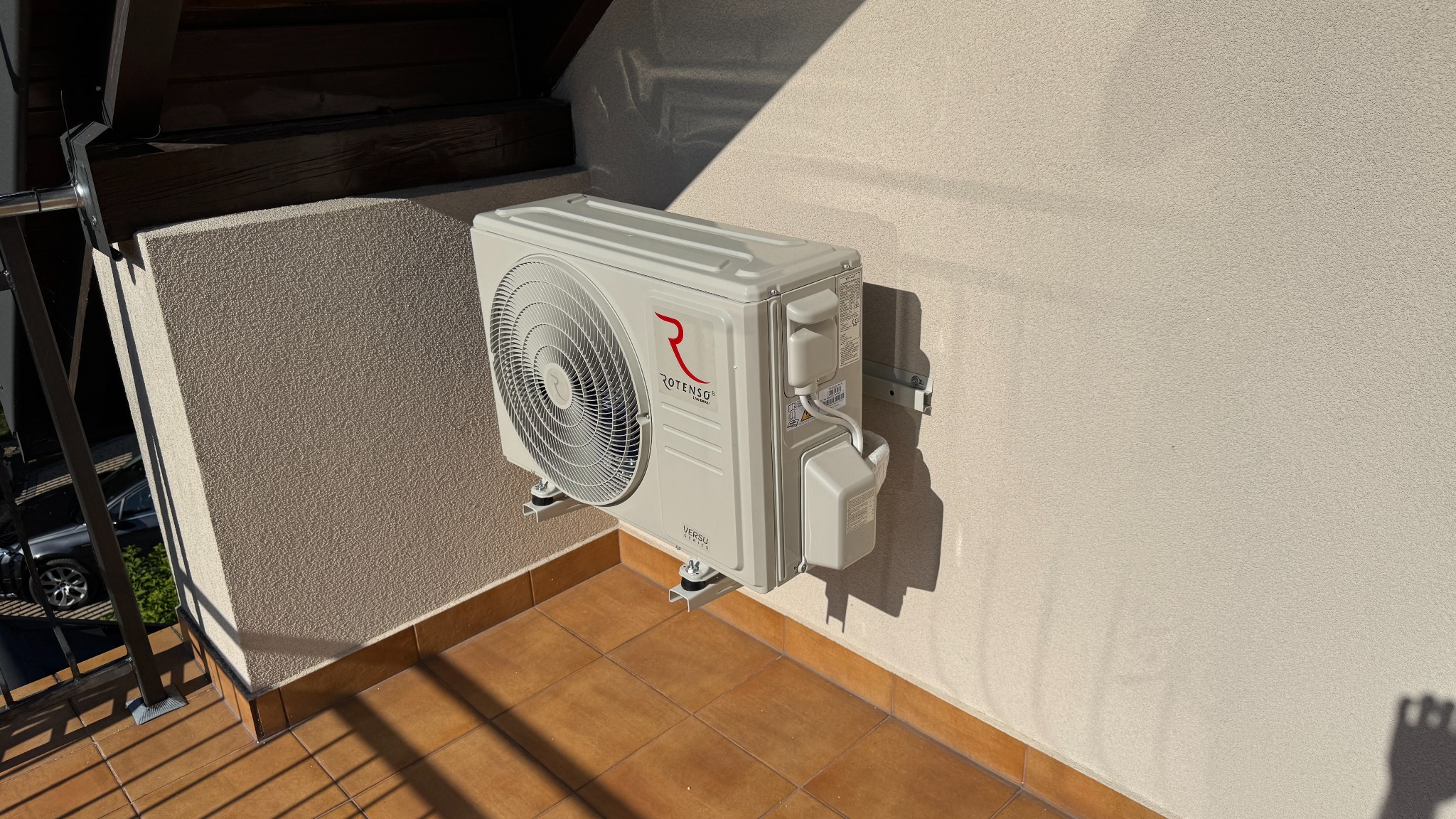 Zewnętrzna jednostka klimatyzacji Rotenso Versu zamontowana na balkonie z terakotą, widoczna z boku, w słoneczny dzień. Metalowe wsporniki.