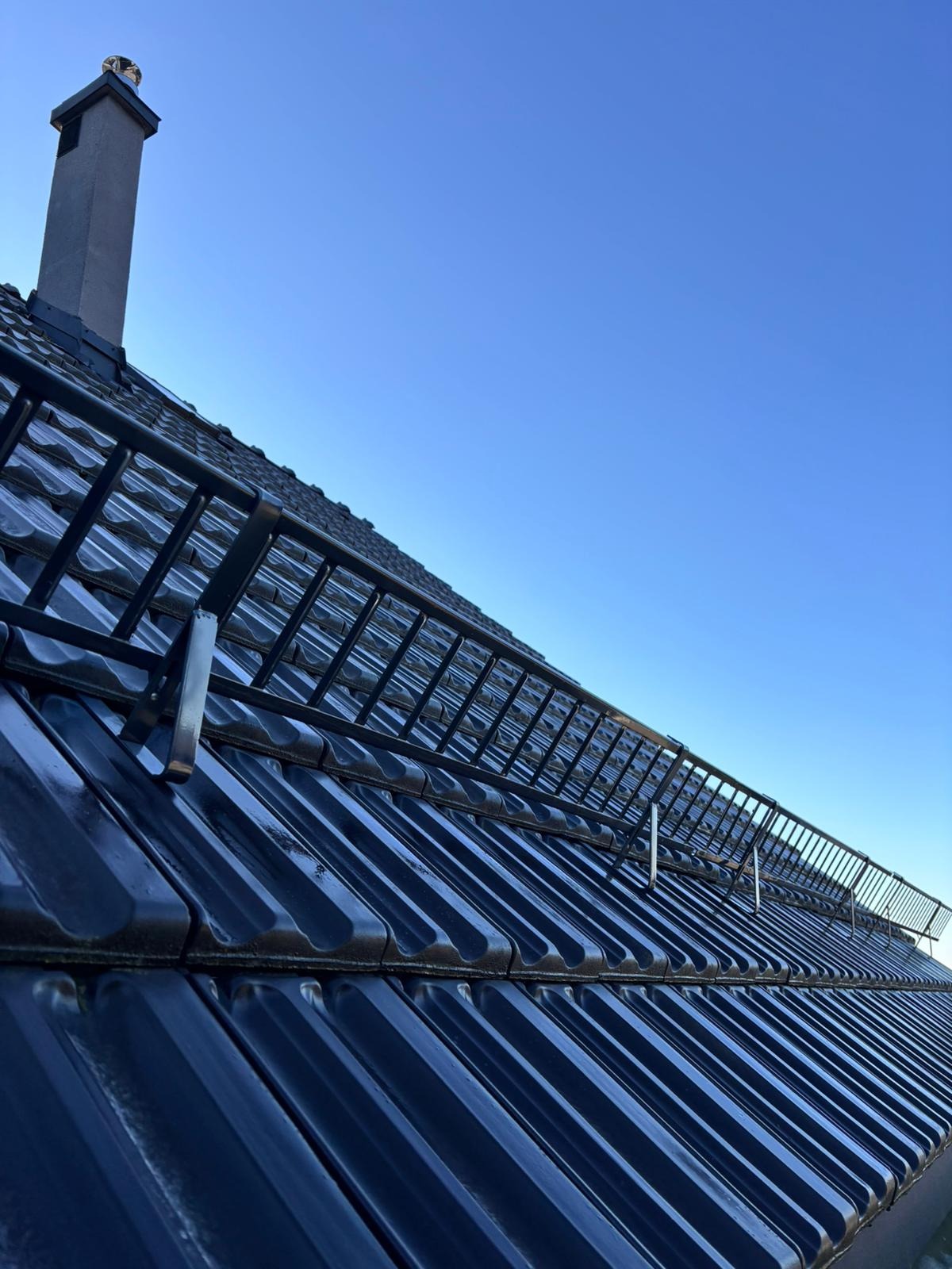 Błyszczący, ciemny dach z zabezpieczeniem przeciwśniegowym i kominem na tle błękitnego nieba, sfotografowany pod kątem z dołu.