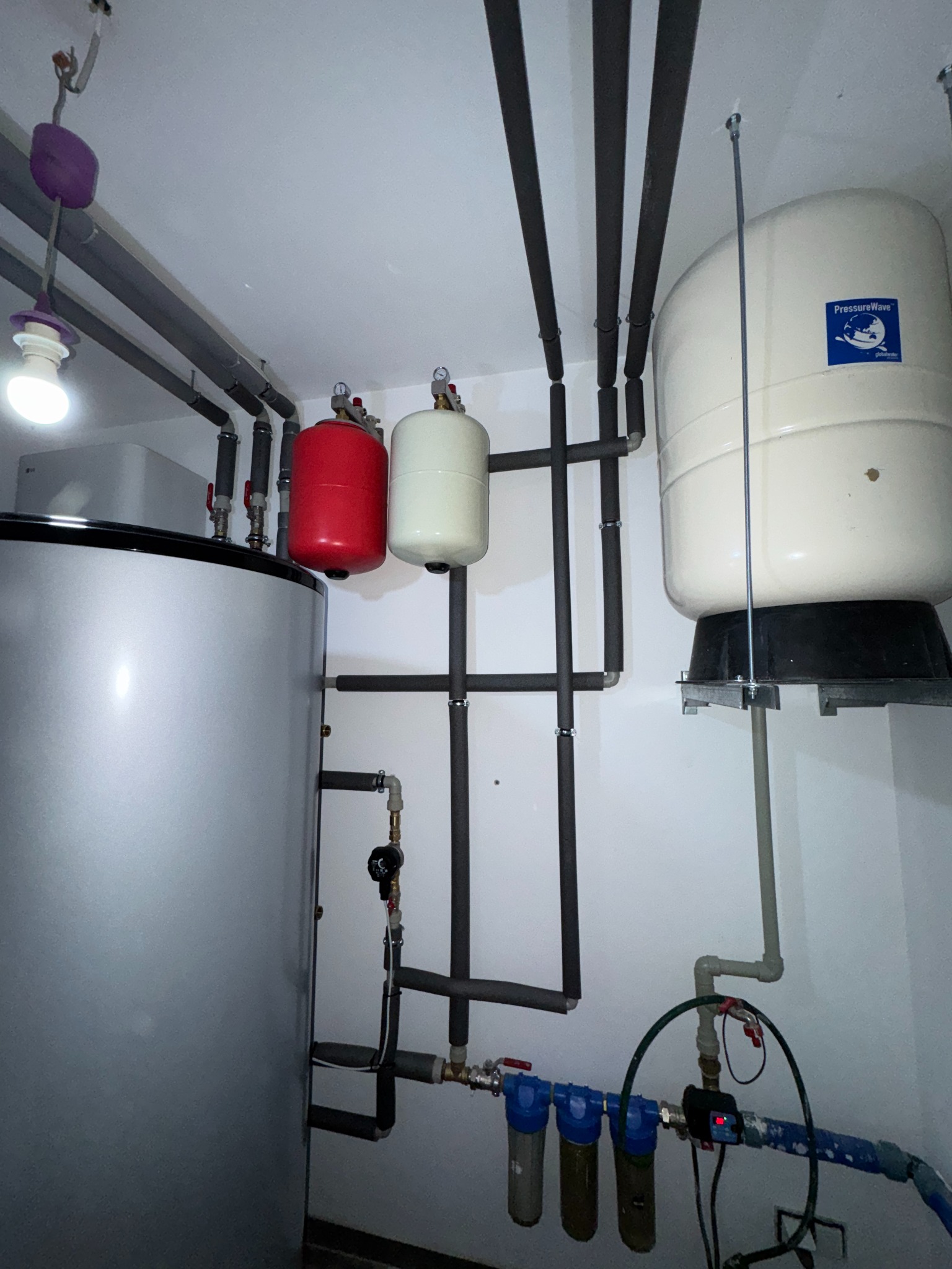 Instalacja CO z szarymi rurami, czerwonym i białym zbiornikiem wyrównawczym, oraz filtrami wody. Widoczne elementy instalacji grzewczej w kotłowni.