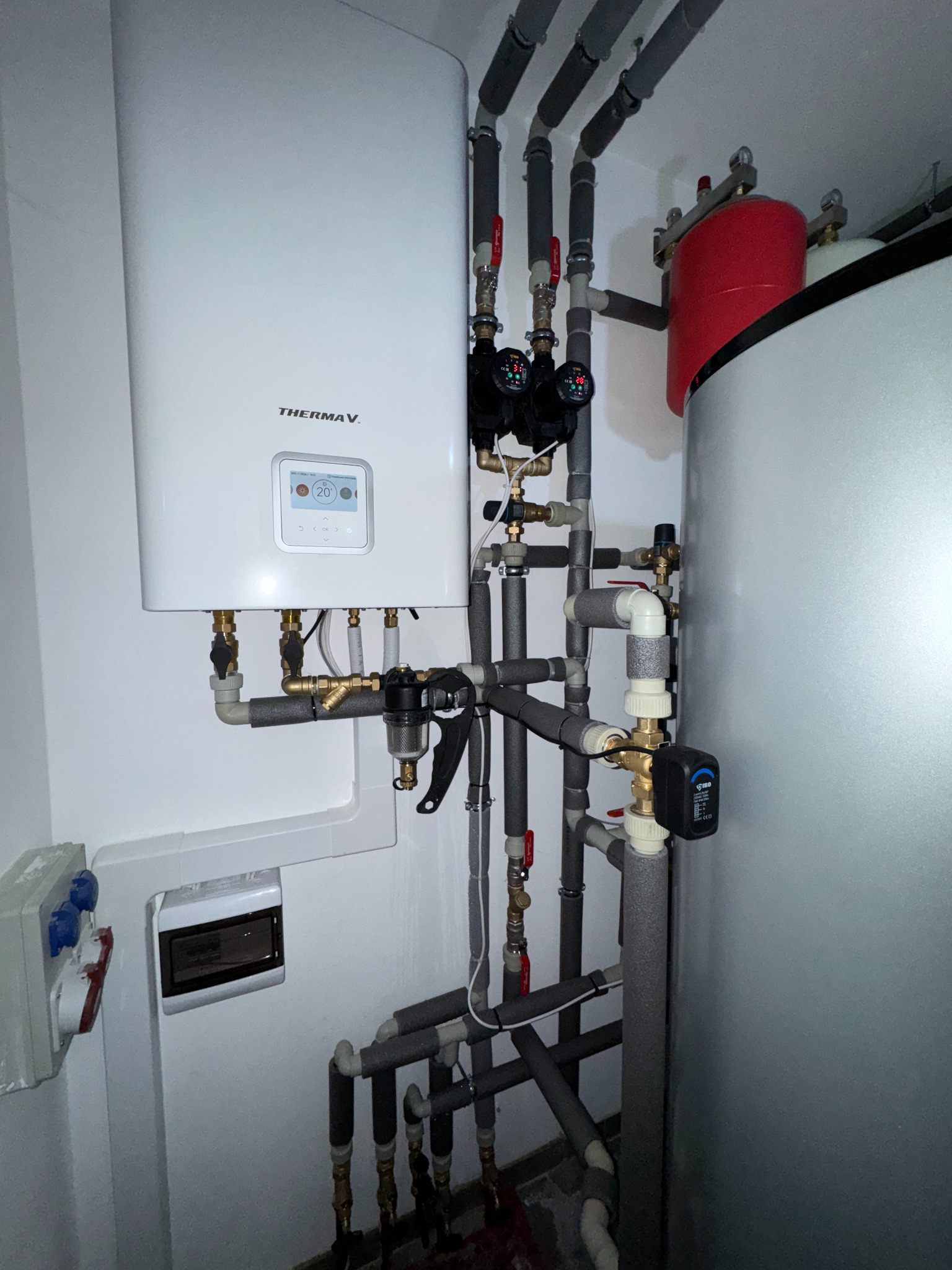 Biały piec grzewczy Therma V z podłączonymi rurami i osprzętem, widoczne zawory, manometry i zbiornik akumulacyjny. Instalacja w pomieszczeniu technicznym.