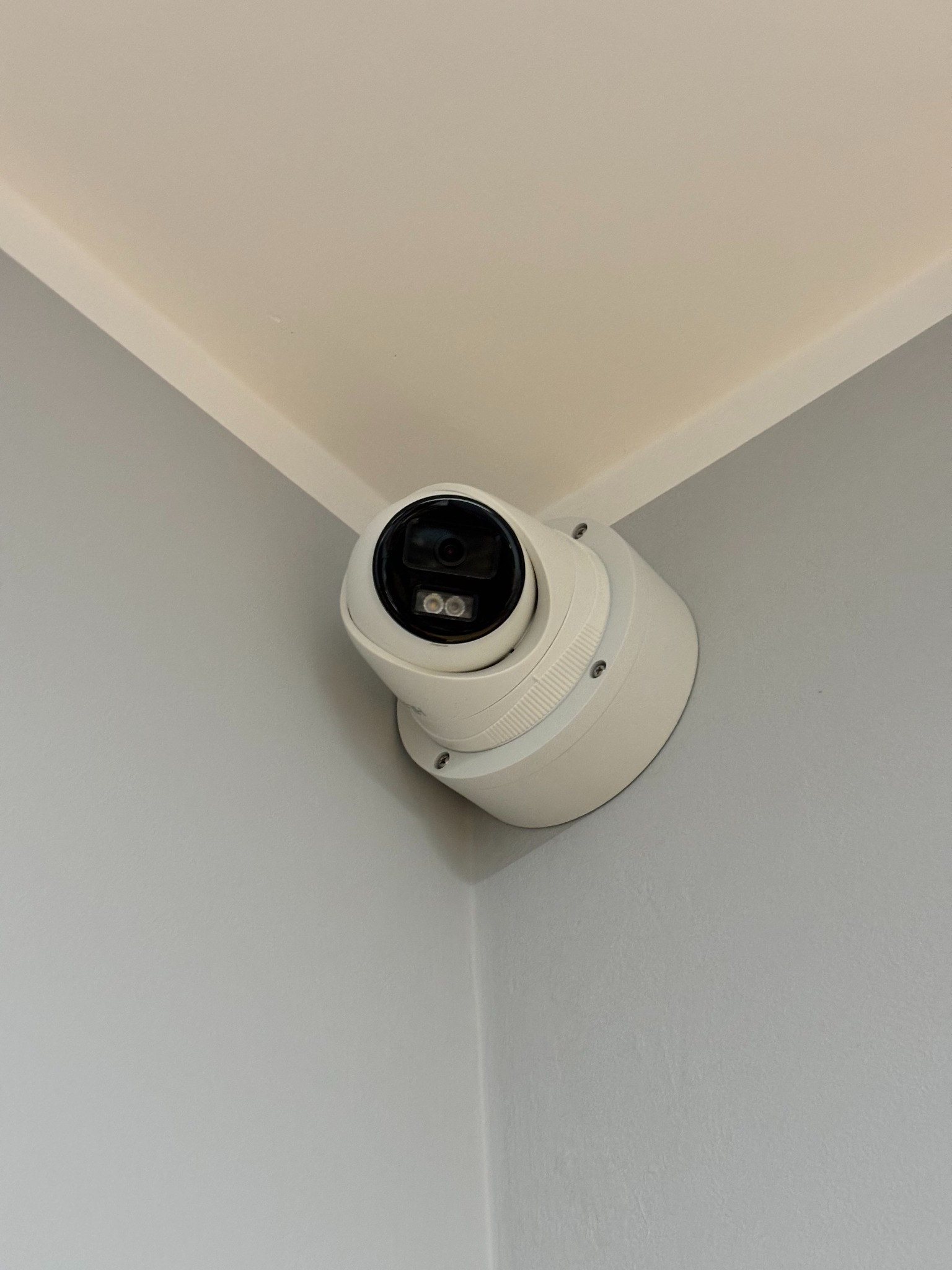 Kamera kopułkowa CCTV zamontowana w rogu pomieszczenia, widok z dołu. Biała obudowa kamery kontrastuje z szarą ścianą i kremowym sufitem.