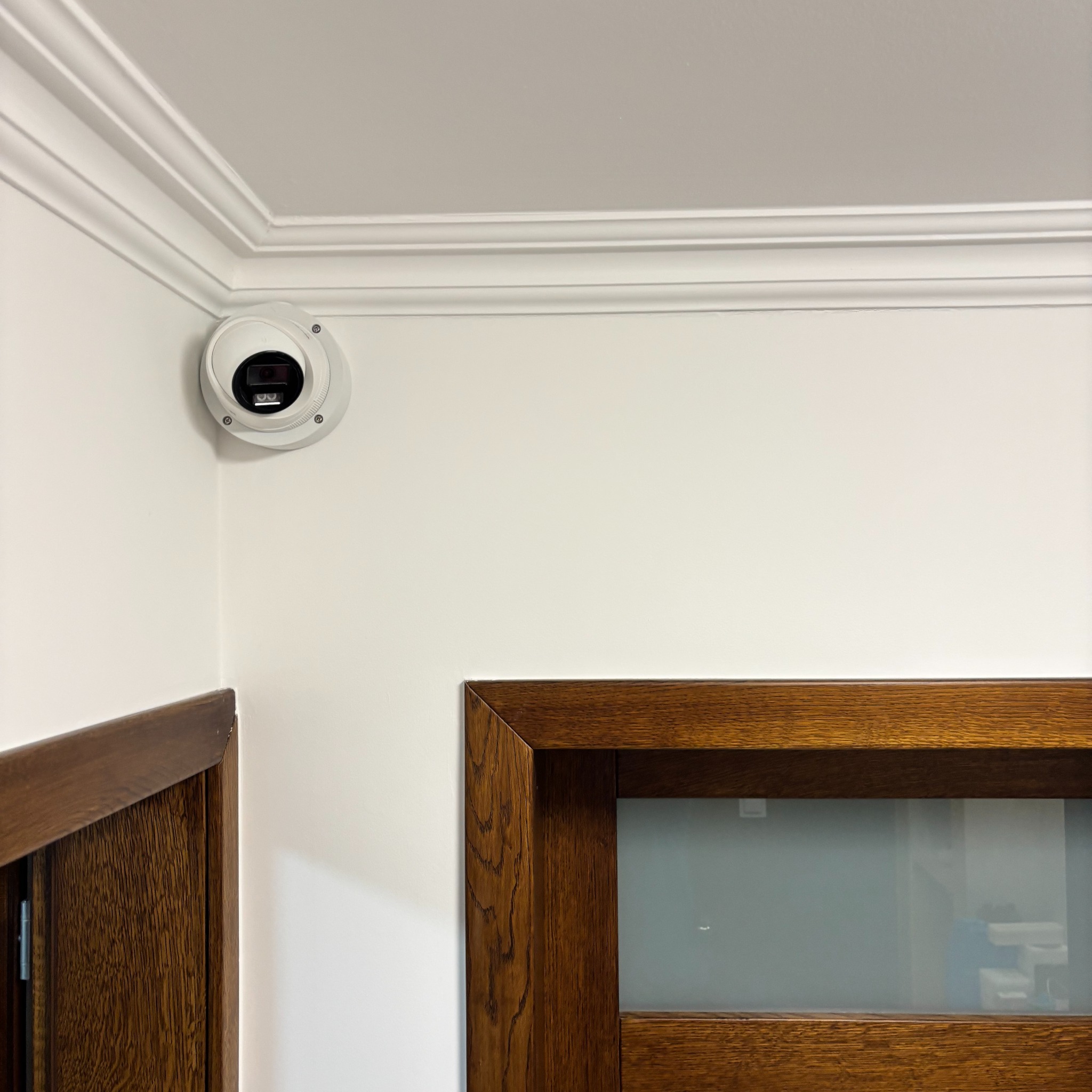 Biała kamera kopułkowa zamontowana w rogu pokoju z dekoracyjną listwą sufitową i fragmentami drewnianych futryn drzwiowych.