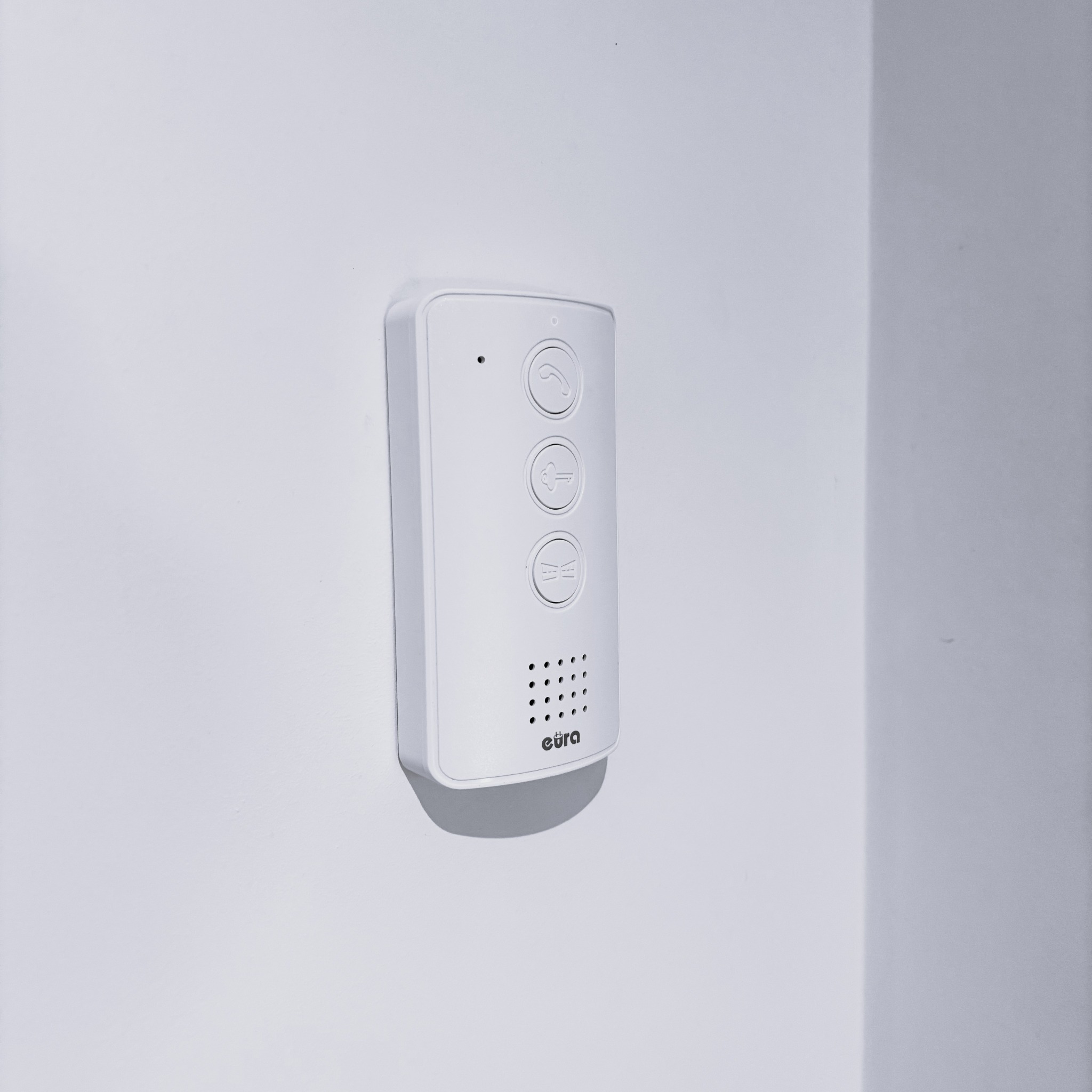 Biały panel domofonu Eura zamontowany na jasnej ścianie, z trzema przyciskami: słuchawka, klucz i głośnik. Minimalistyczny design, czyste tło.