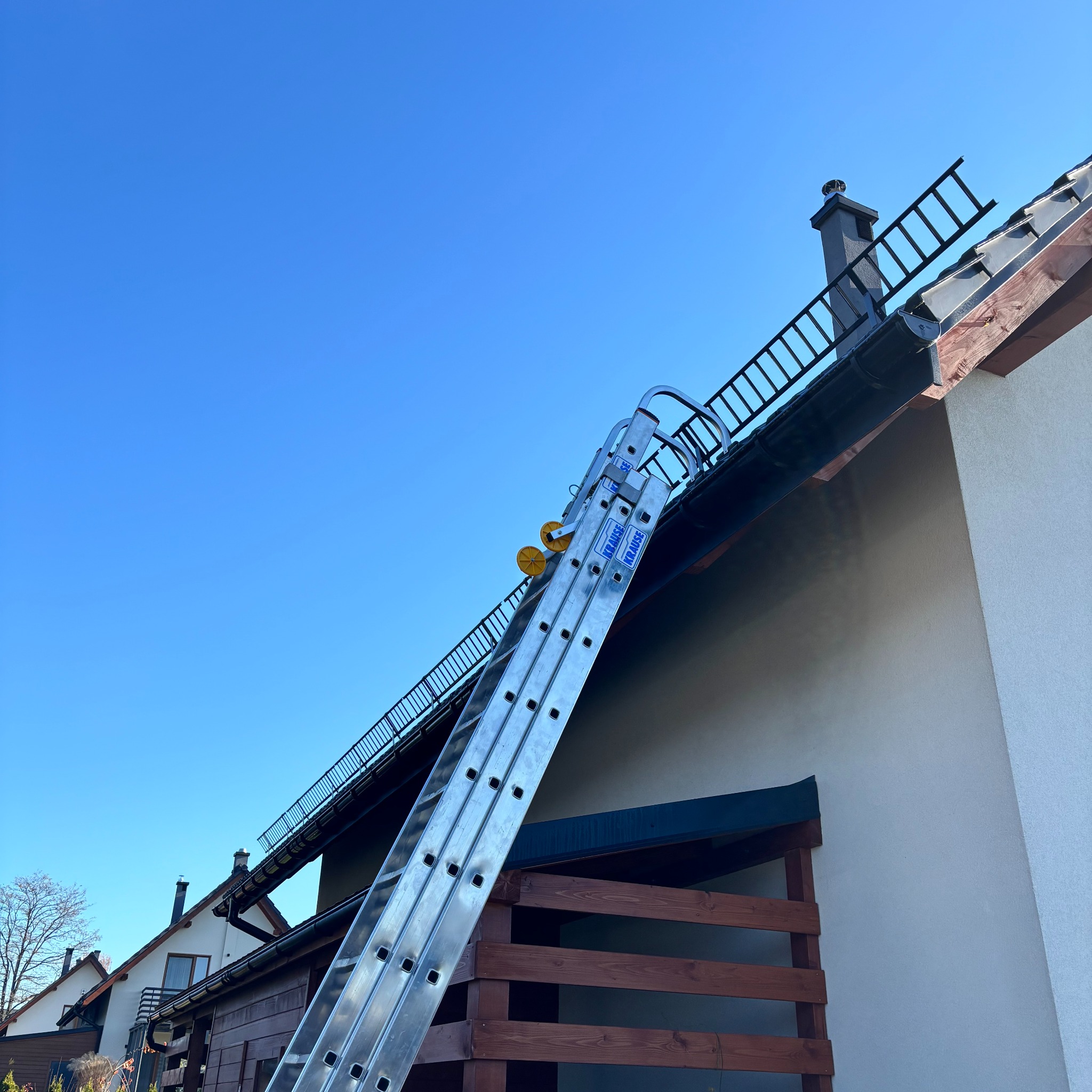 Aluminiowa drabina oparta o dach domu jednorodzinnego, z widocznym kominem i rynną. Błękitne niebo w tle. Prace na wysokościach.